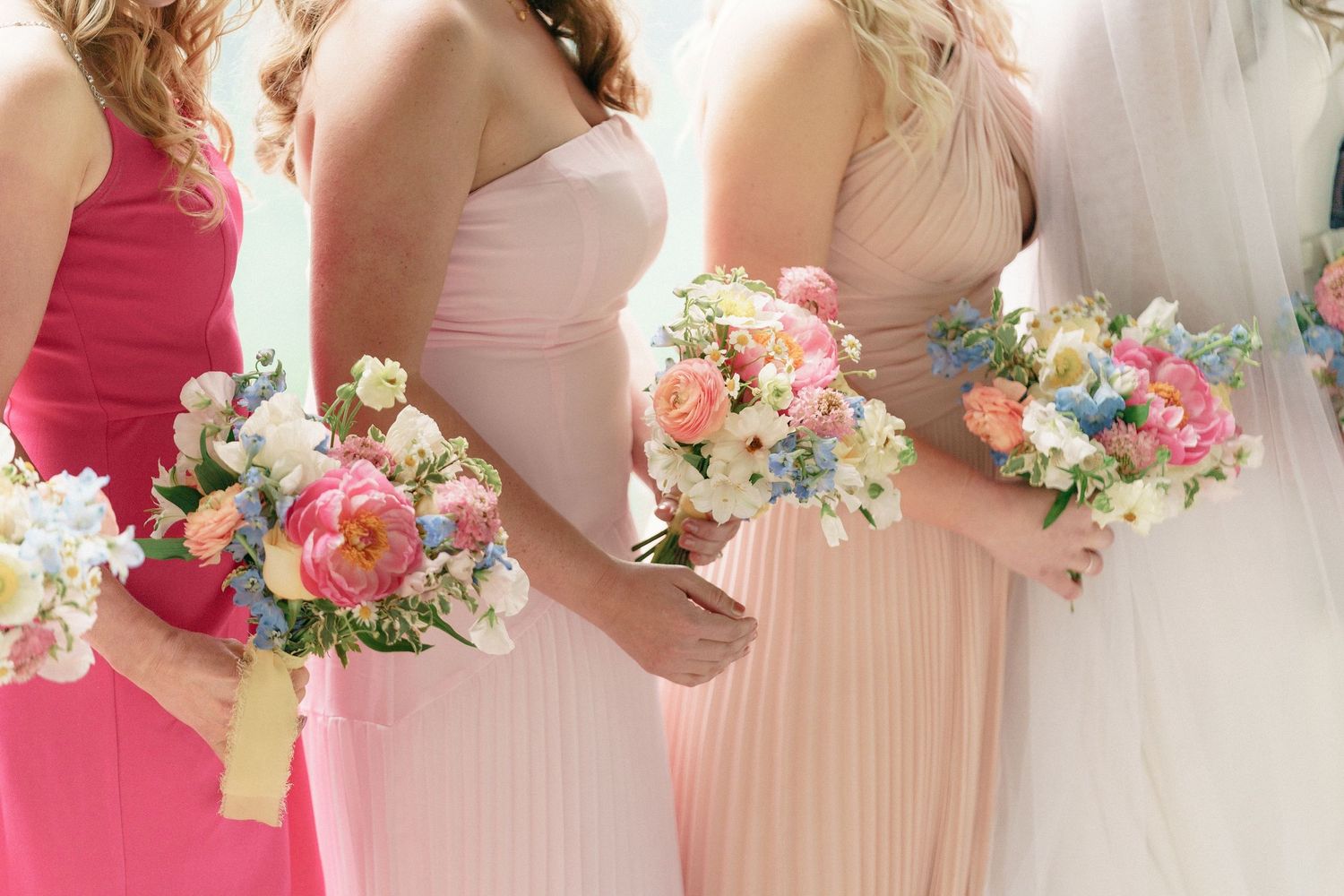 A wedding party stands together holding pink and white floral bouquets while wearing light pink and fuchsia dresses.