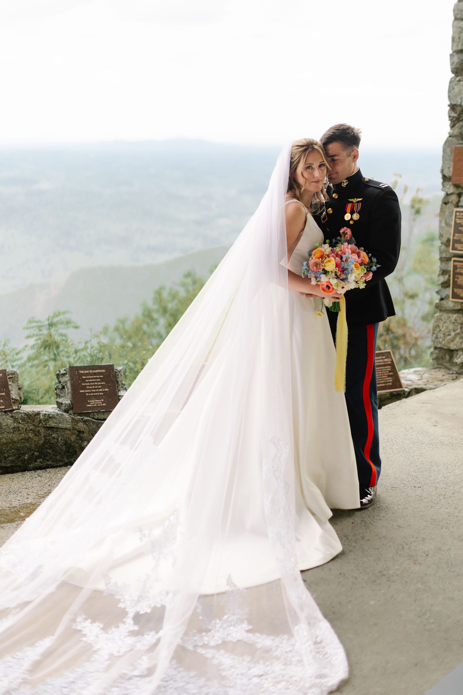 Military wedding couple embraces on scenic mountain overlook with flowing white veil and colorful bouquet.
