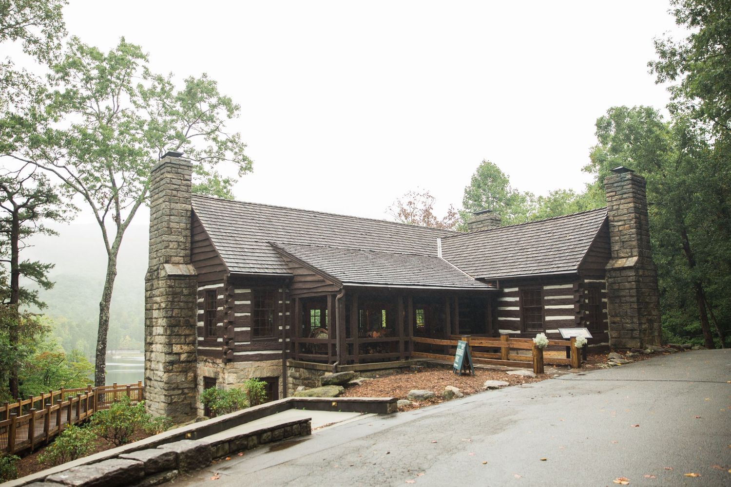 Rustic log cabin with stone chimneys and wooden porches sits on a misty hillside surrounded by green trees.