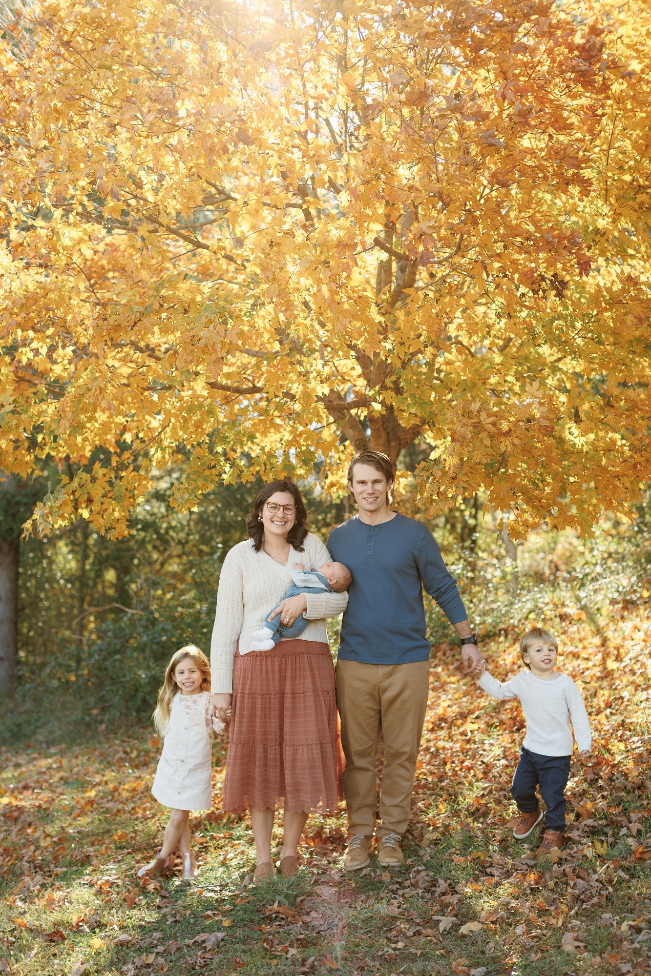 Family of four sits on blanket under golden autumn tree, parents holding newborn while children play with falling leaves.