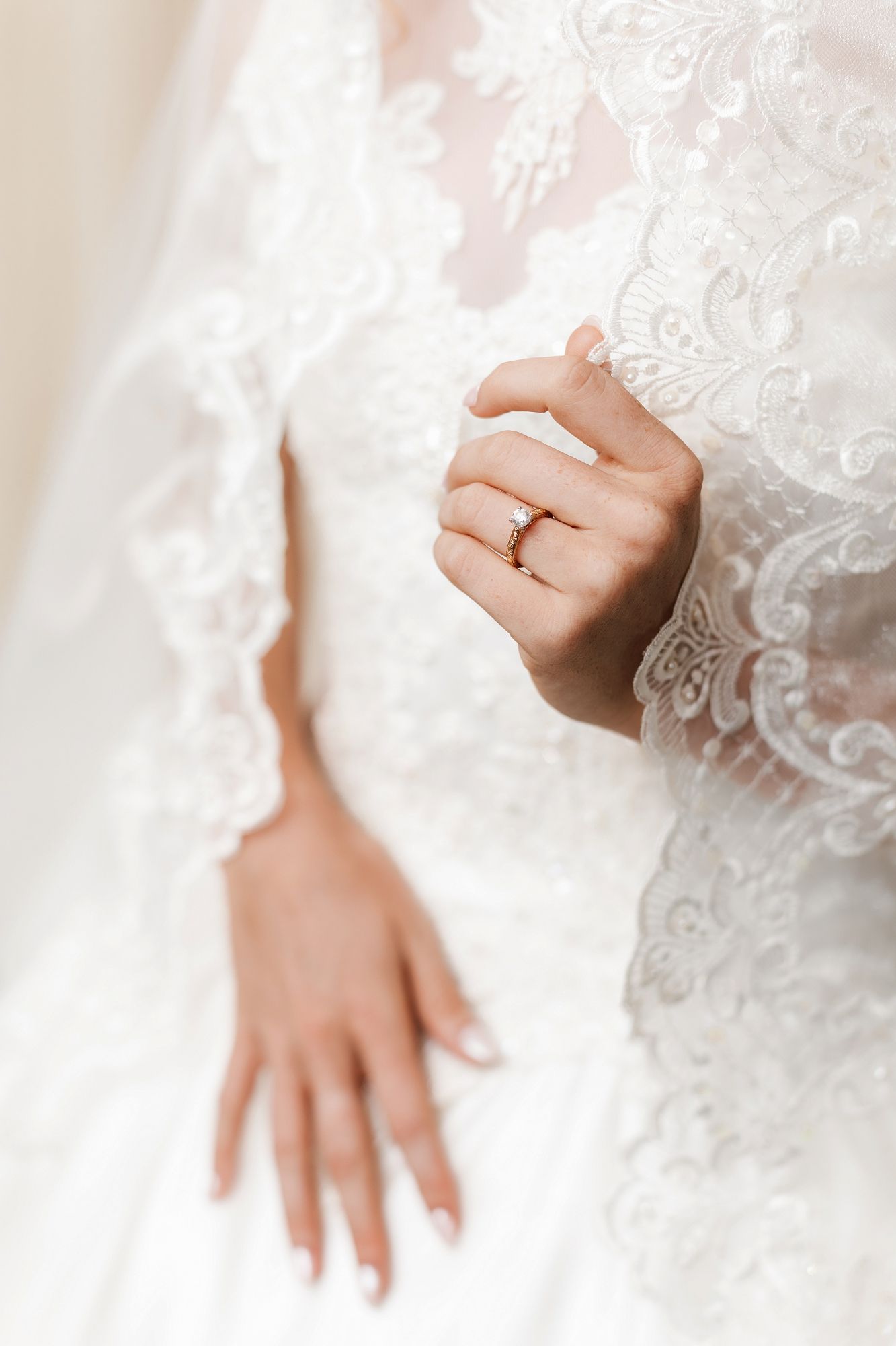 Close-up of a bride's hands showing an elegant diamond engagement ring on her finger against a lace wedding dress.