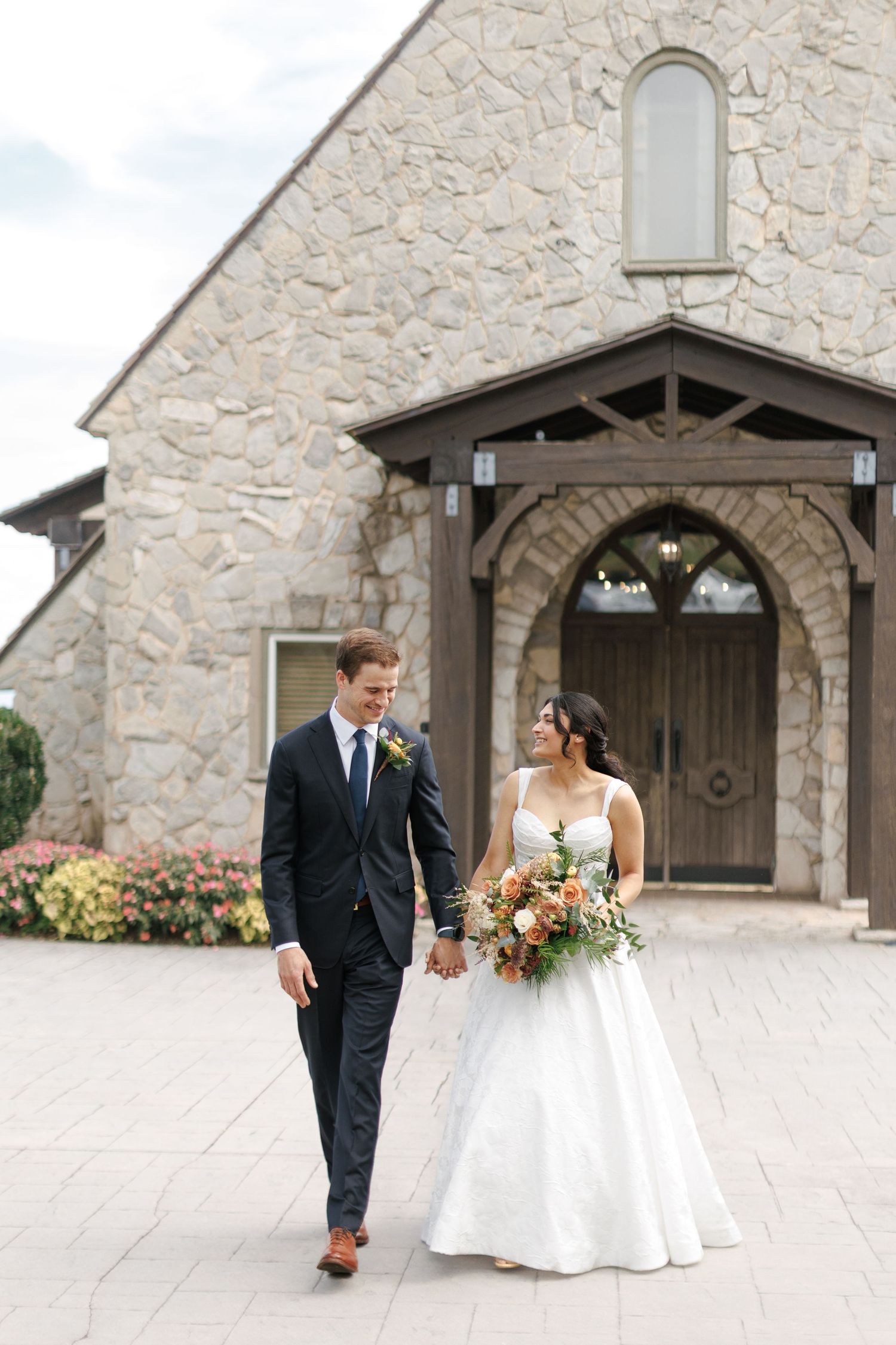 Bride and groom walk hand-in-hand before a stone chapel, she holds a lush bouquet in a white ballgown.