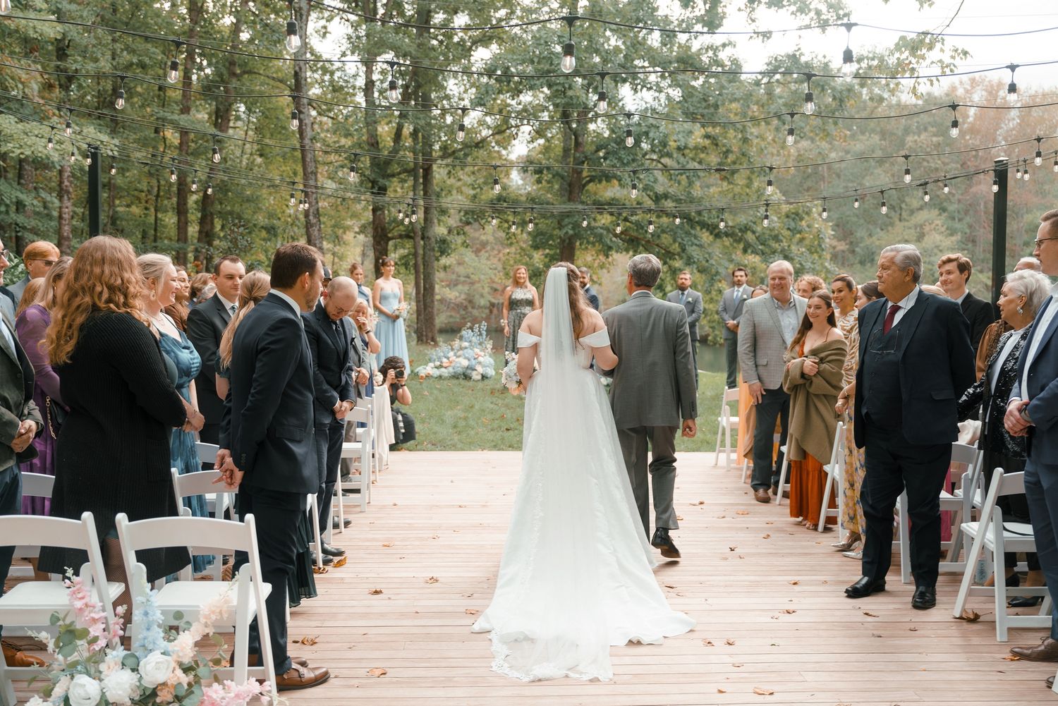Bride walks down outdoor aisle with escort at woodland wedding ceremony with string lights.