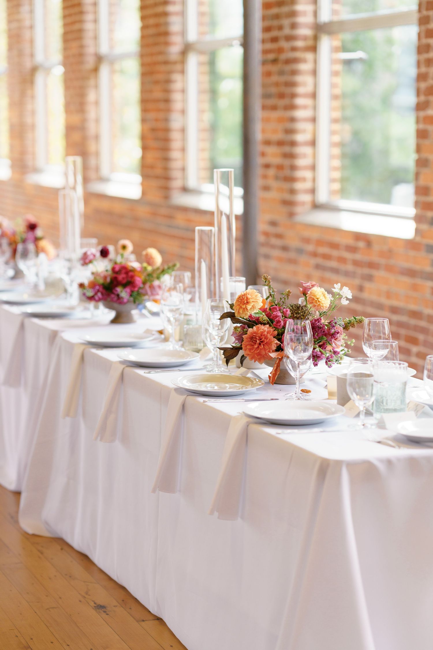Elegant wedding reception table with white linen, orange floral centerpieces, tall candles, and wine glasses in a brick venue.