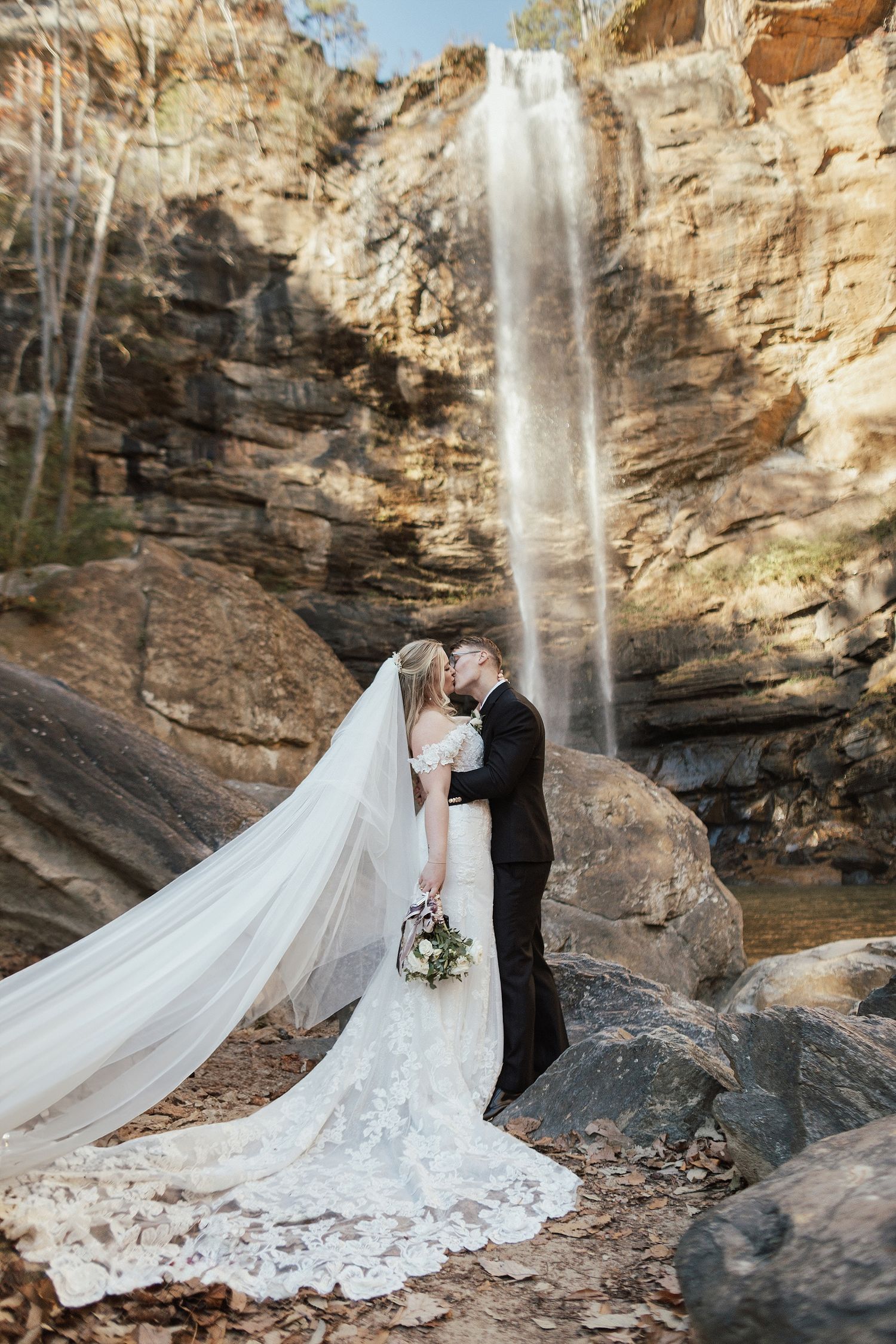 A couple shares an intimate moment in front of a majestic waterfall, with the bride wearing a flowing lace gown with long veil.