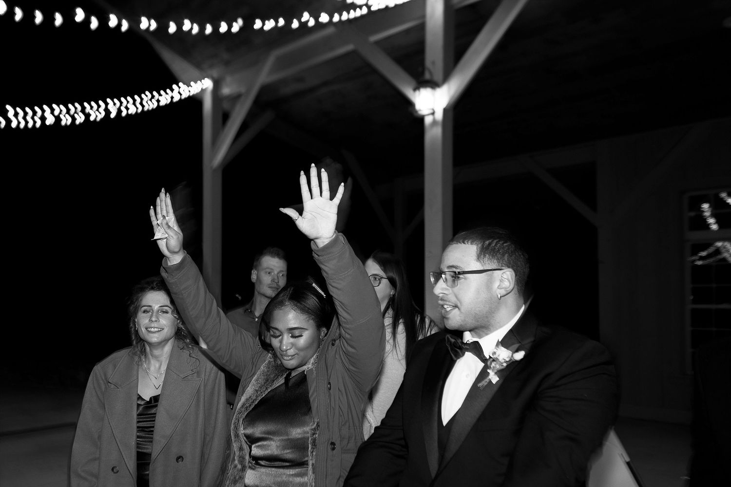 Black and white photo of people celebrating with raised hands under string lights at an evening event.