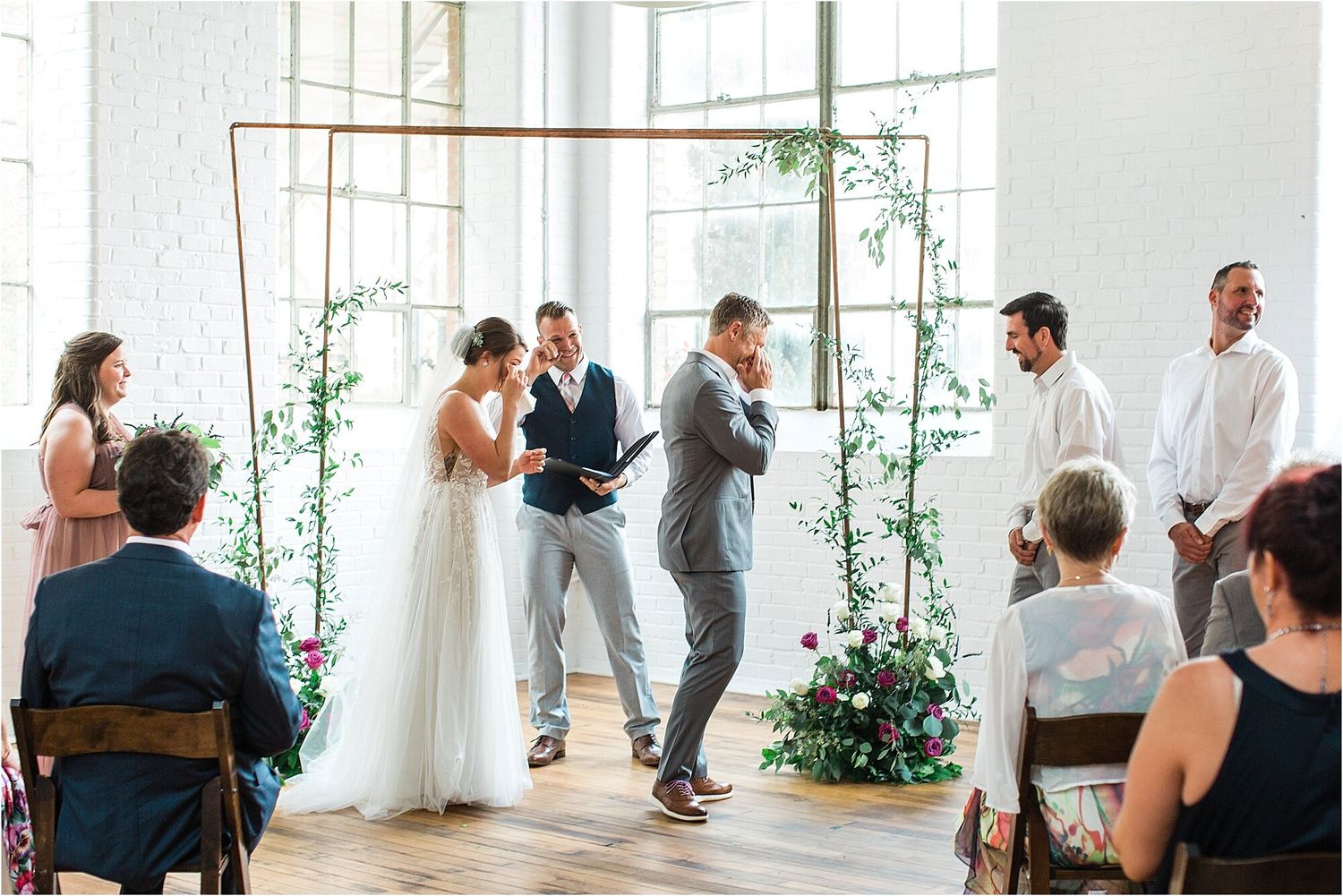 Wedding ceremony in a modern white venue with copper arch and greenery decorations.