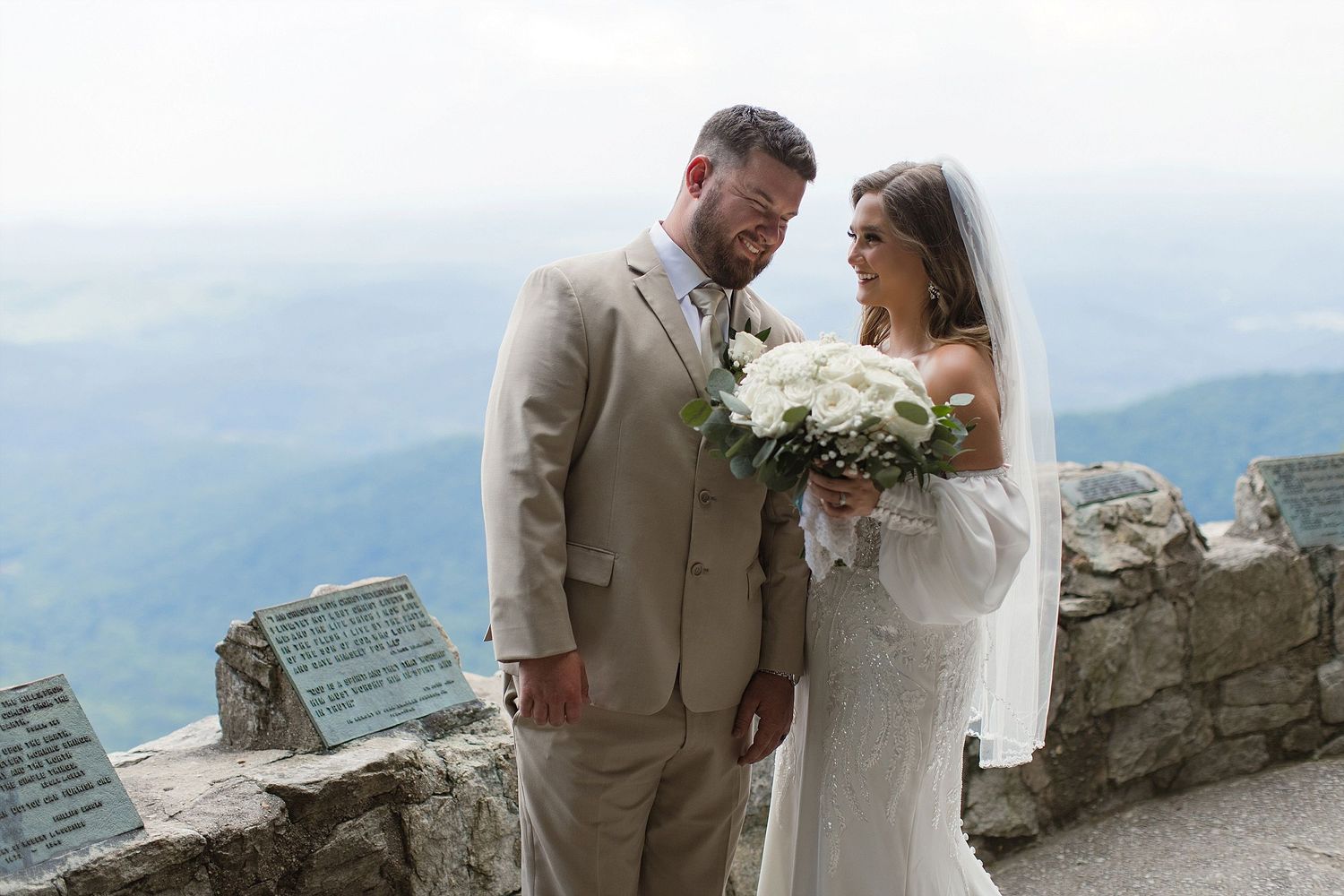 Newlyweds share an intimate moment on a stone overlook with mountain views in the background.