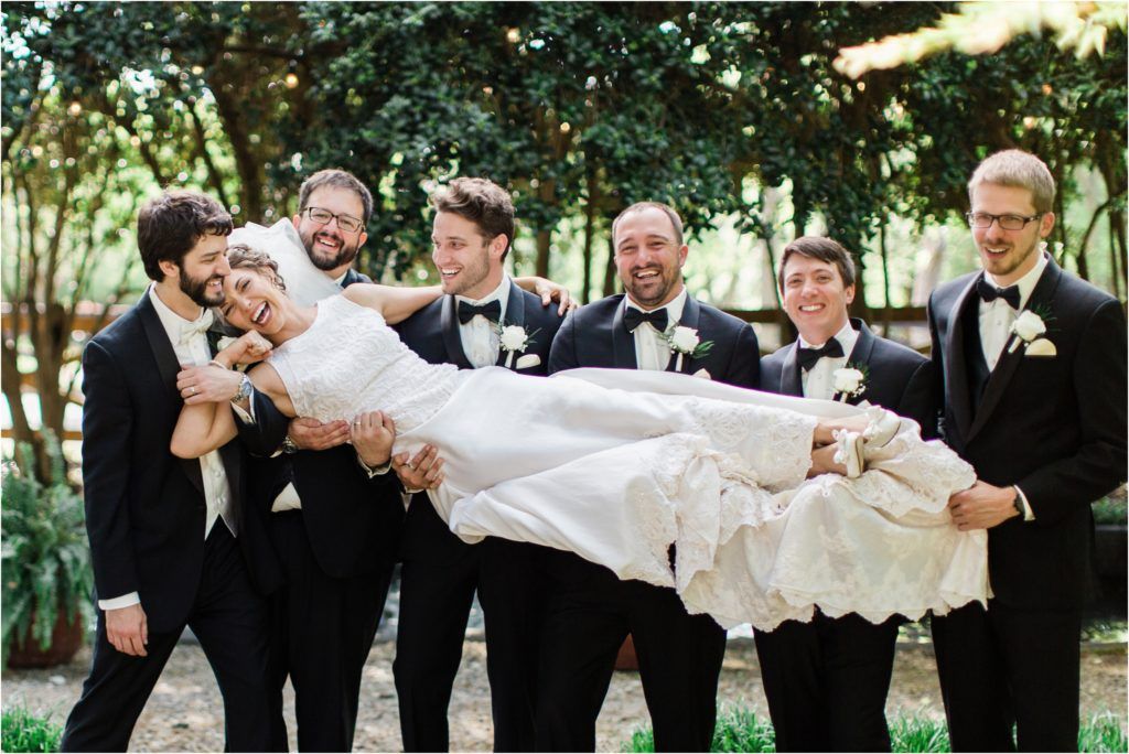 Groomsmen in black tuxedos playfully carrying bride in white wedding dress outdoors.