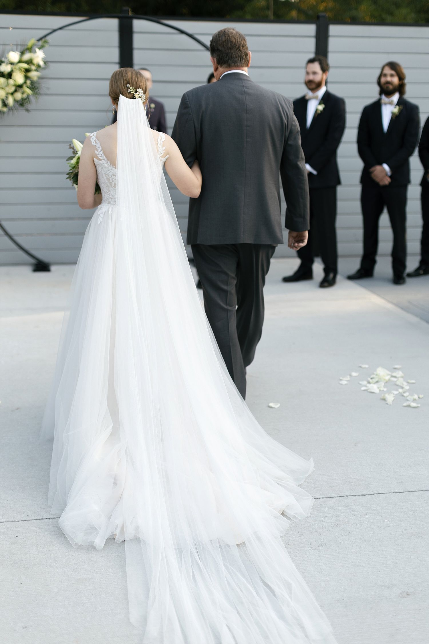 A bride in a flowing white gown with a long train walks down the aisle with someone in a dark suit during an outdoor ceremony.