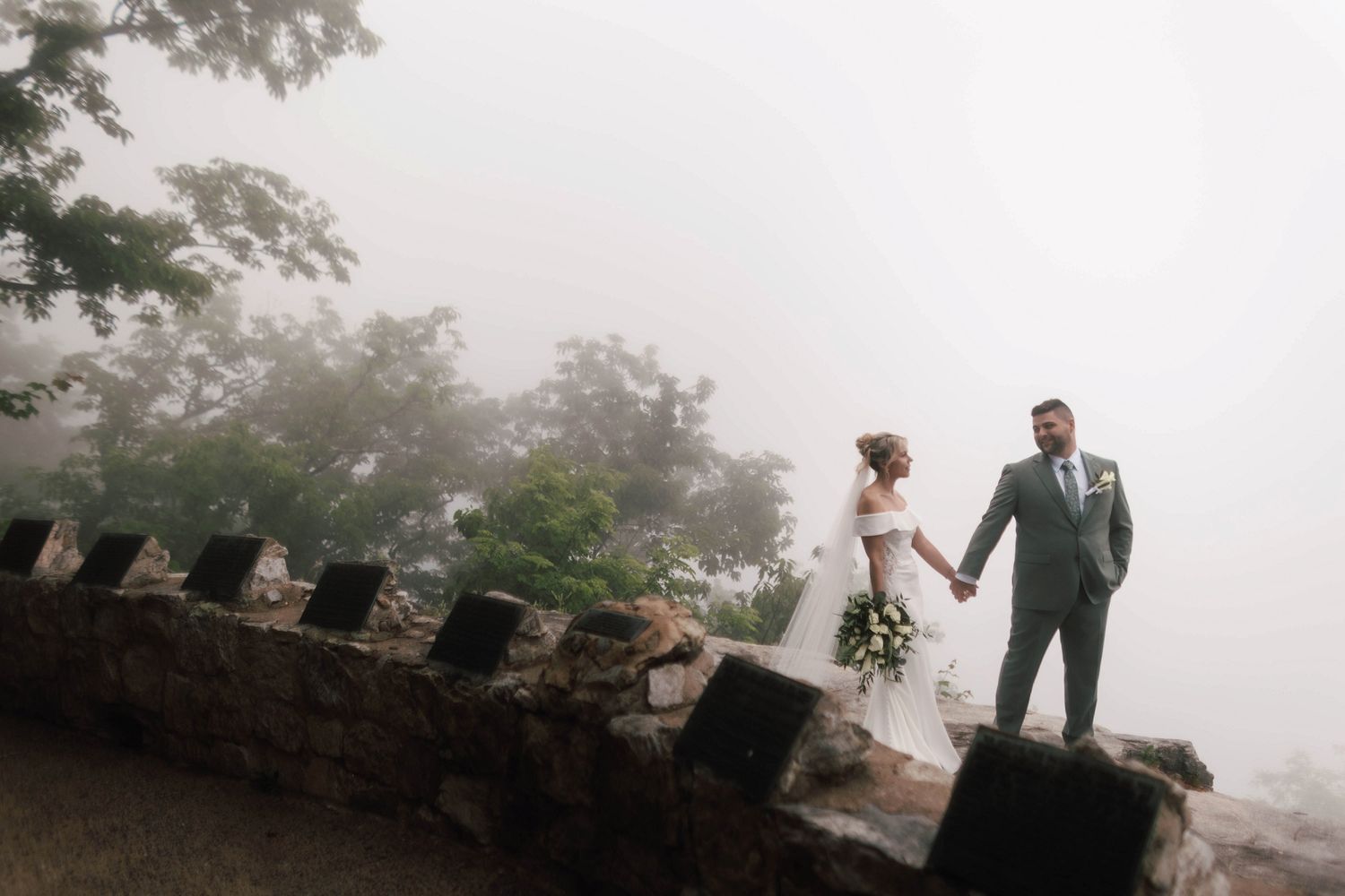 Bride and groom holding hands walking along a stone wall overlook surrounded by fog and trees.