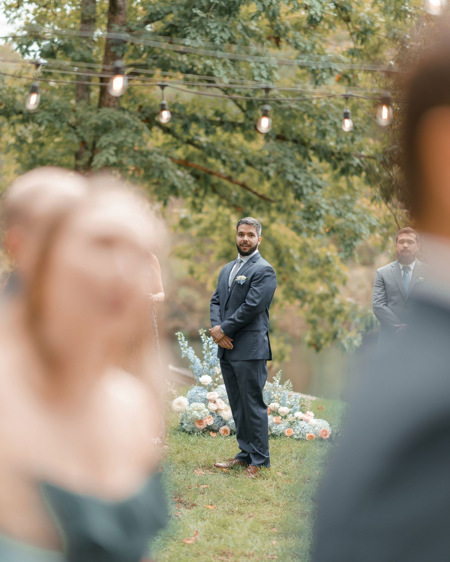 Groom in navy suit awaits bride at outdoor wedding ceremony with string lights and floral arrangements.