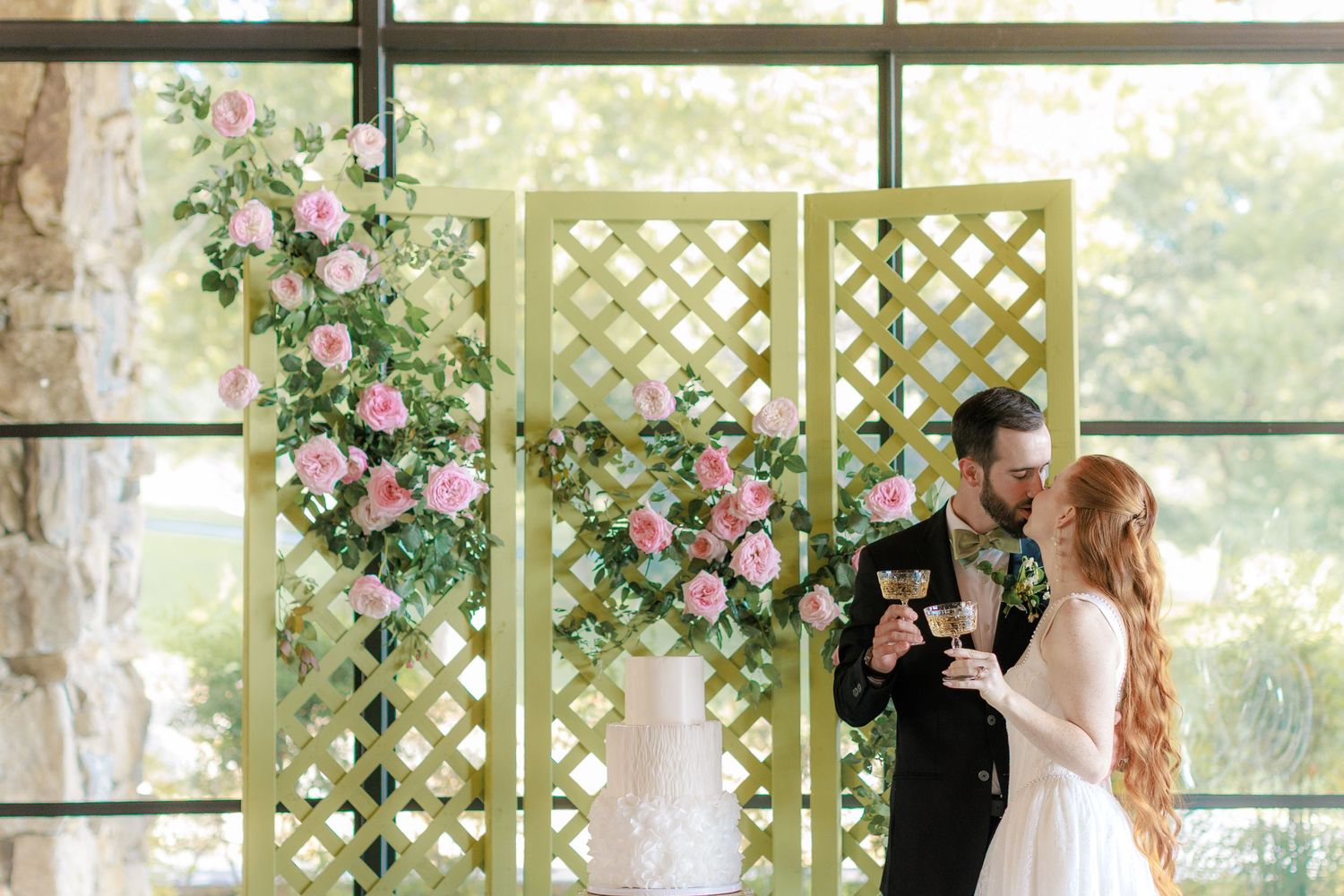 Bride and groom toasting with wine glasses beside a yellow lattice backdrop adorned with pink roses and a wedding cake.