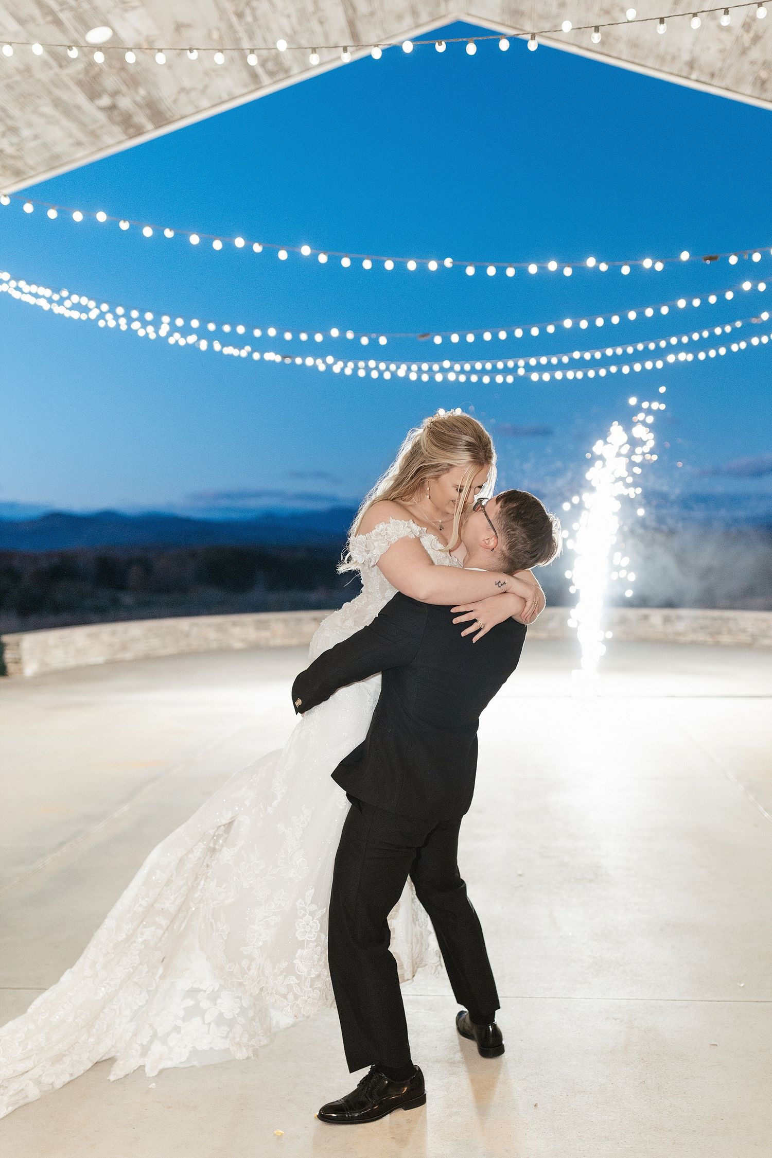 A couple dances under twinkling string lights at an outdoor wedding reception with mountains visible in the background.