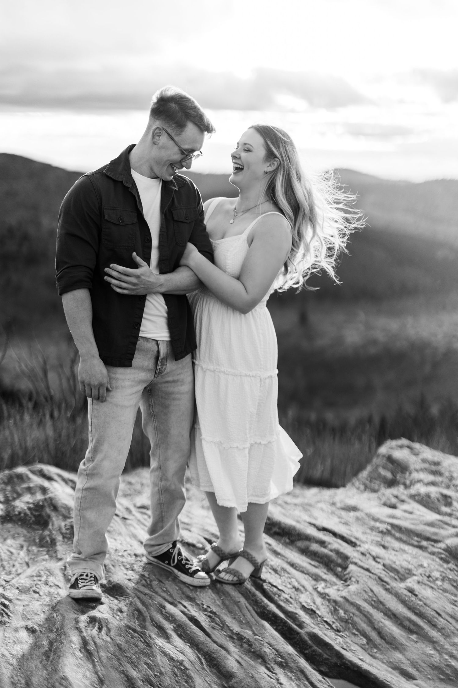 A couple shares a joyful candid moment together while standing on a rocky mountain overlook in black and white.