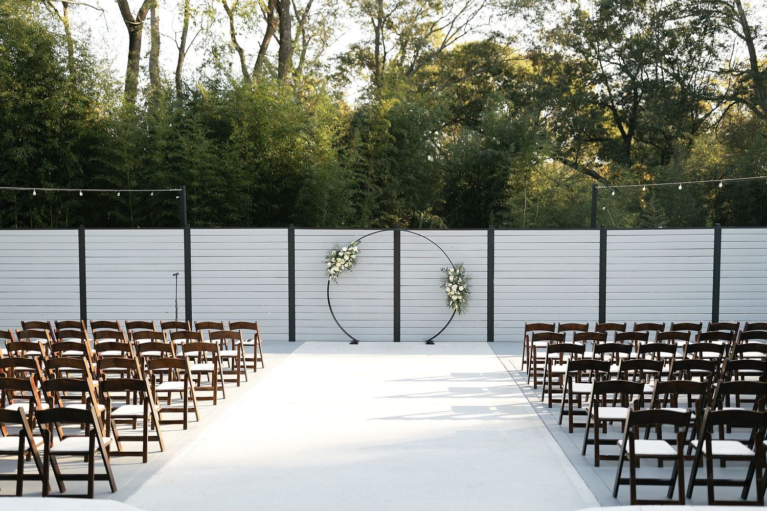 Modern outdoor wedding ceremony setup with wooden chairs and white backdrop wall surrounded by trees.