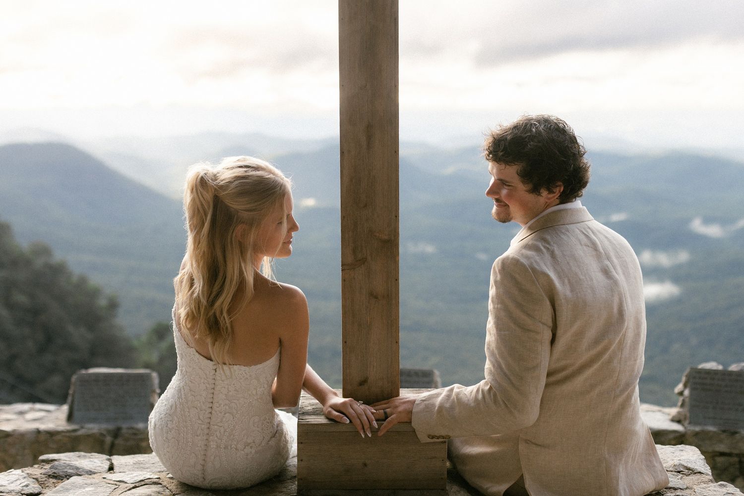 A couple sits together on a stone wall overlooking a scenic mountain landscape on a cloudy day.