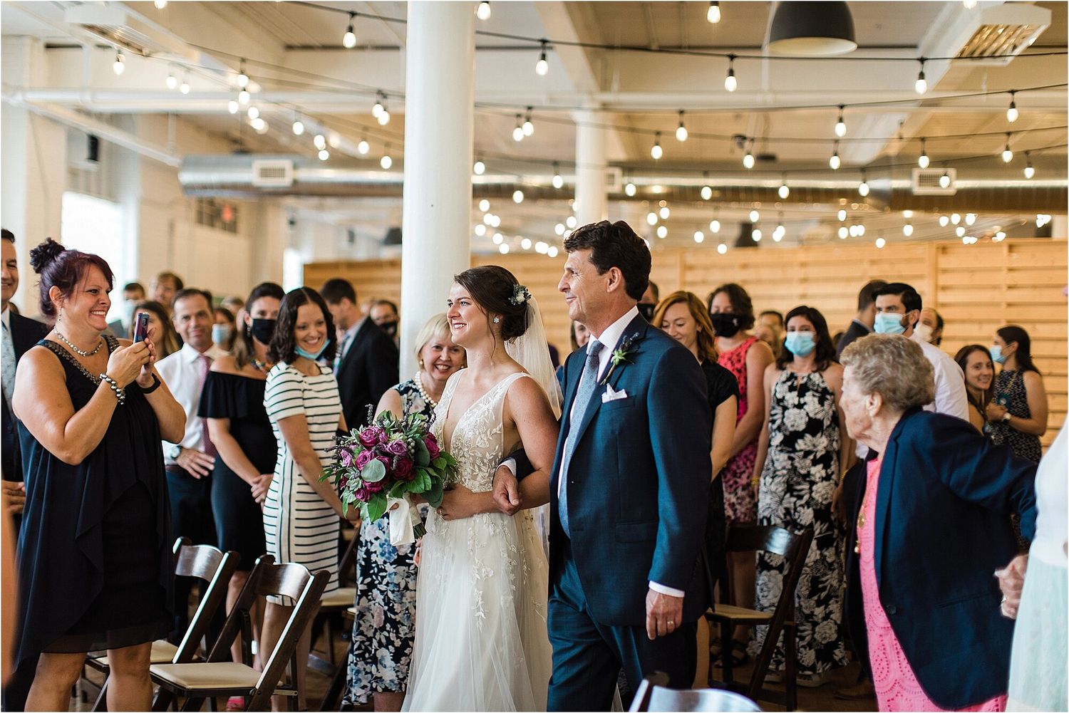 Wedding guests gather in an industrial-style venue with exposed ceiling and string lights during a ceremony.