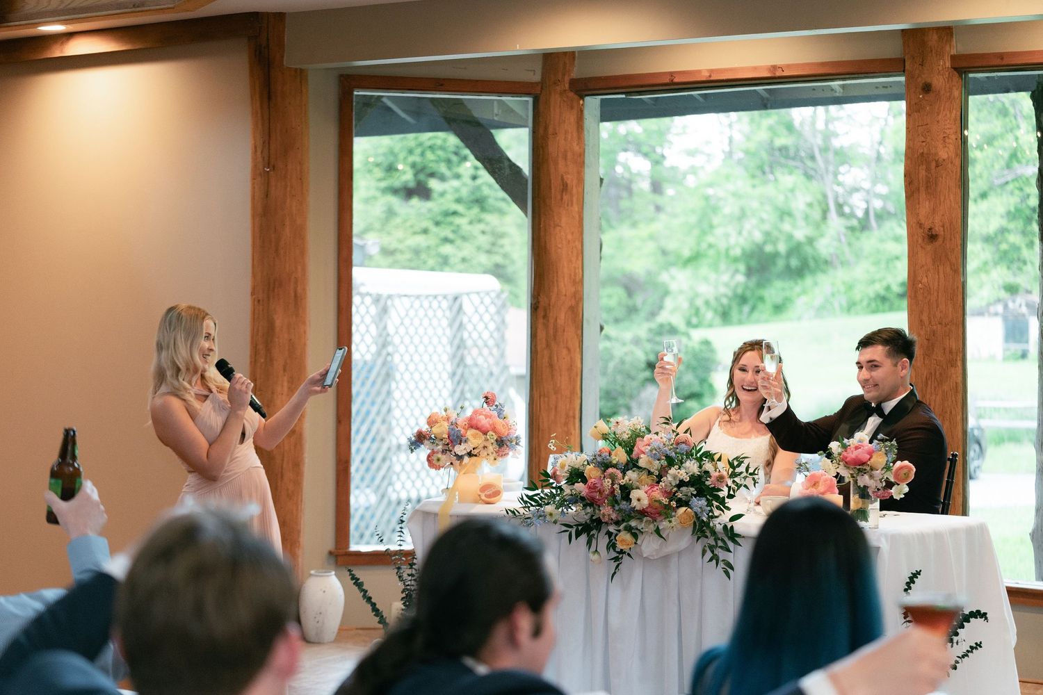 Wedding reception speeches taking place in a bright venue with wood beams and large windows overlooking greenery.