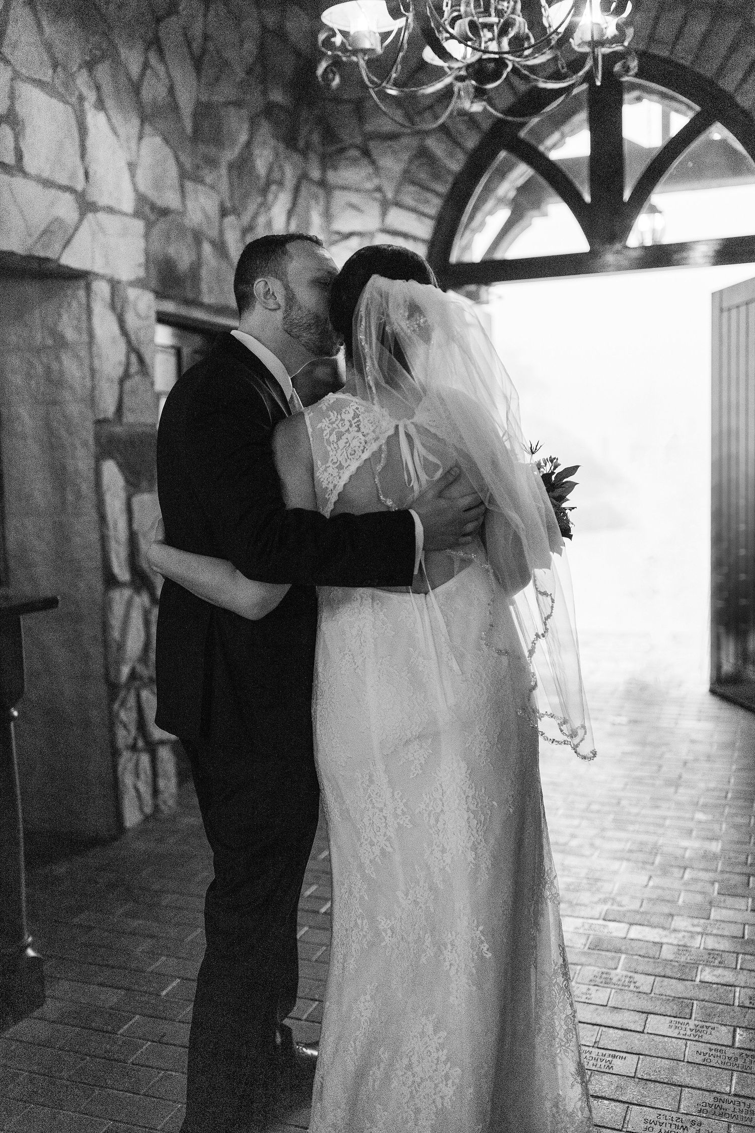 Black and white photo of couple sharing intimate moment in stone hallway with chandelier.