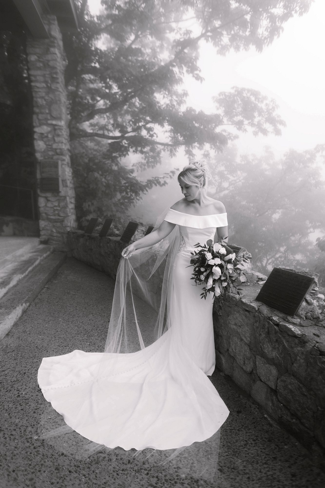 Black and white photo of a bride in an off-shoulder gown holding a bouquet and veil in misty outdoor setting.