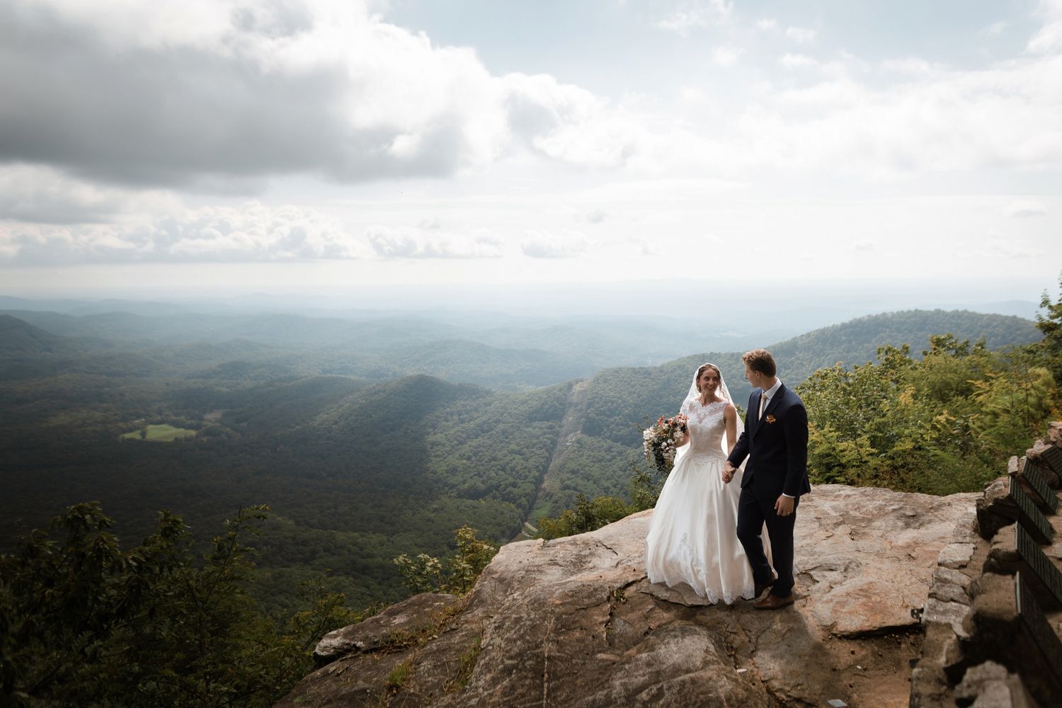 Bride and groom stand on rocky mountain overlook in wedding attire with sweeping green valley views.