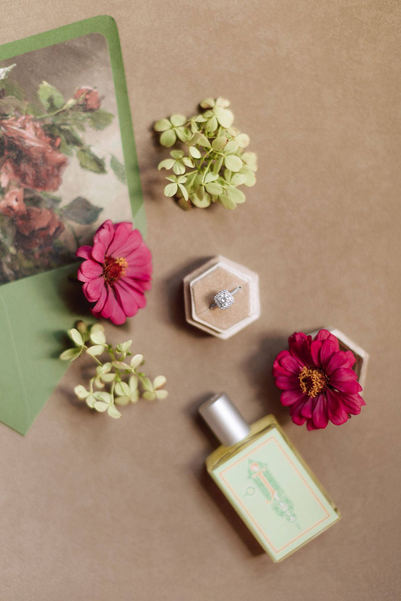 Flat lay of pink zinnias, green foliage, diamond ring in velvet box, perfume bottle, and floral card on tan background.