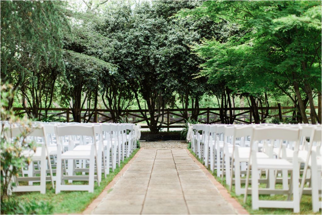 Garden wedding ceremony setup with rows of white chairs along a stone pathway surrounded by lush greenery.