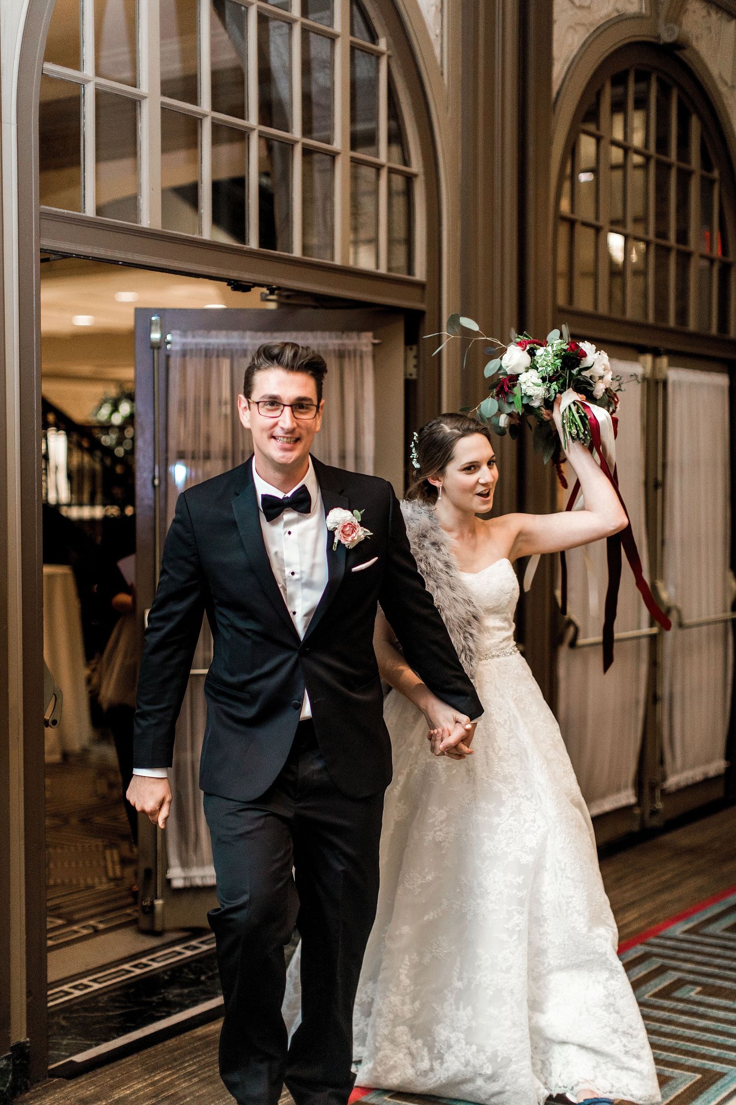 A joyful wedding couple exits through an arched doorway with the bride holding up her bouquet.
