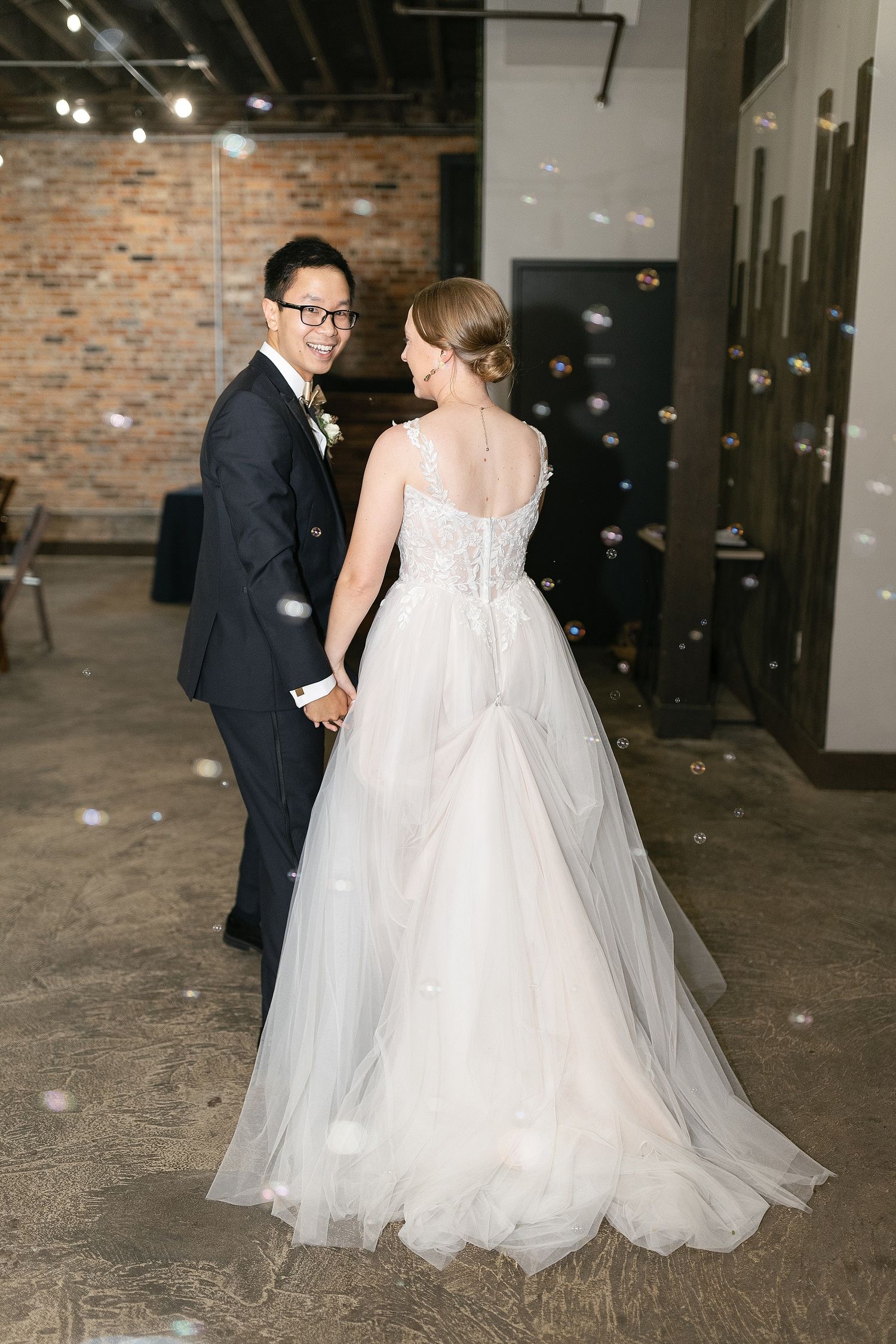 A bride in a flowing white tulle gown and groom in navy suit share a romantic moment in an industrial venue space.