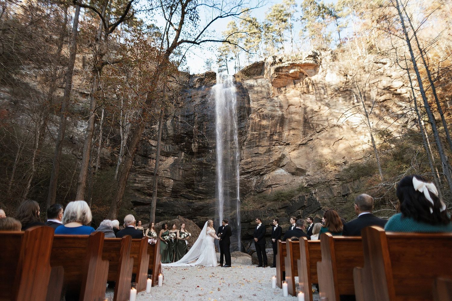 A stunning outdoor wedding ceremony setup with wooden pews facing a majestic waterfall cascading down rocky cliffs.