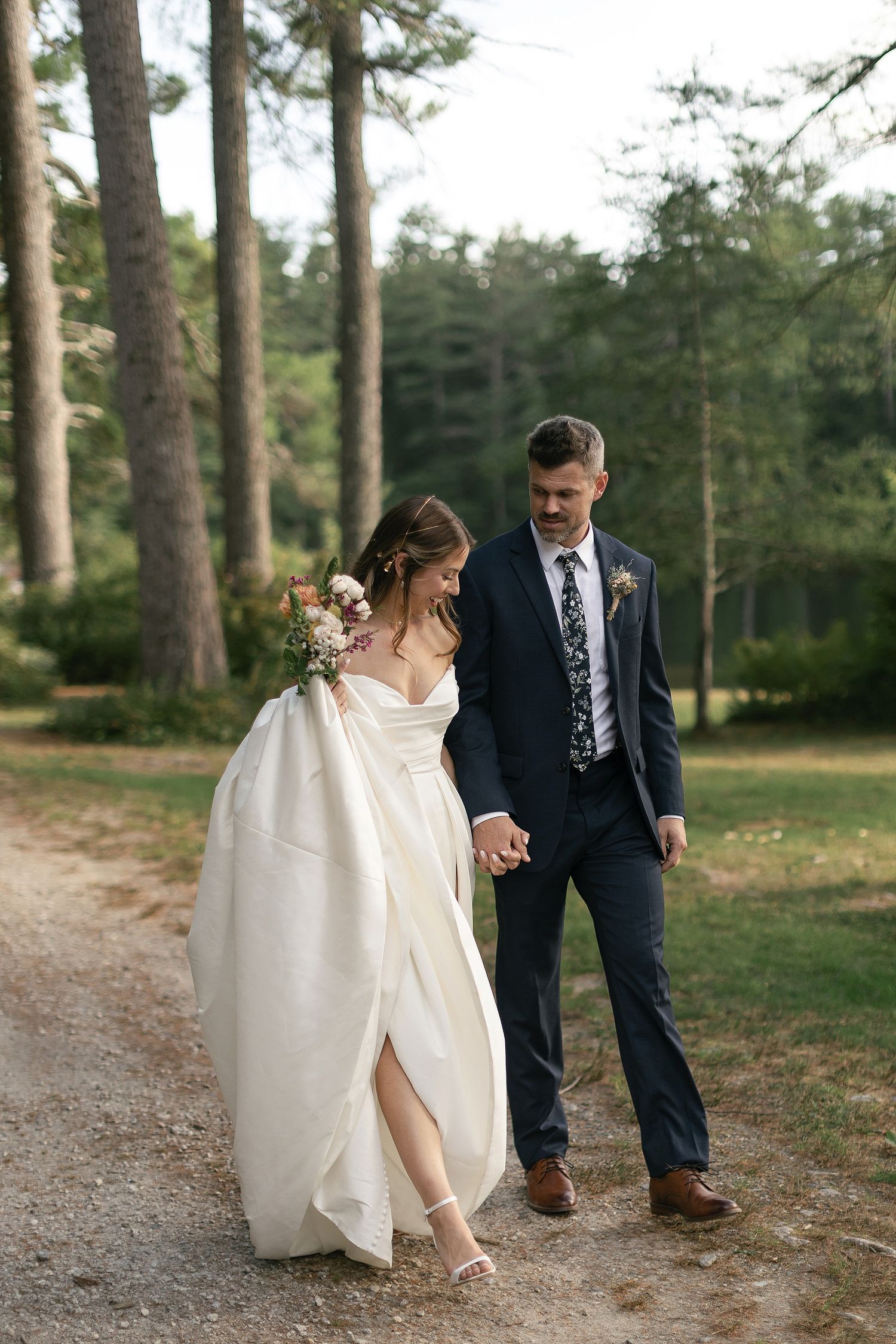 A couple walks hand in hand along a forest path in wedding attire.