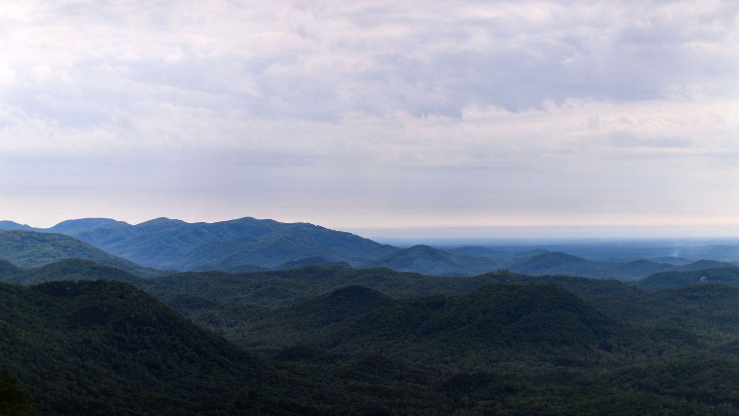 A panoramic view of rolling mountains with layers of forested peaks stretching to the misty horizon under cloudy skies.