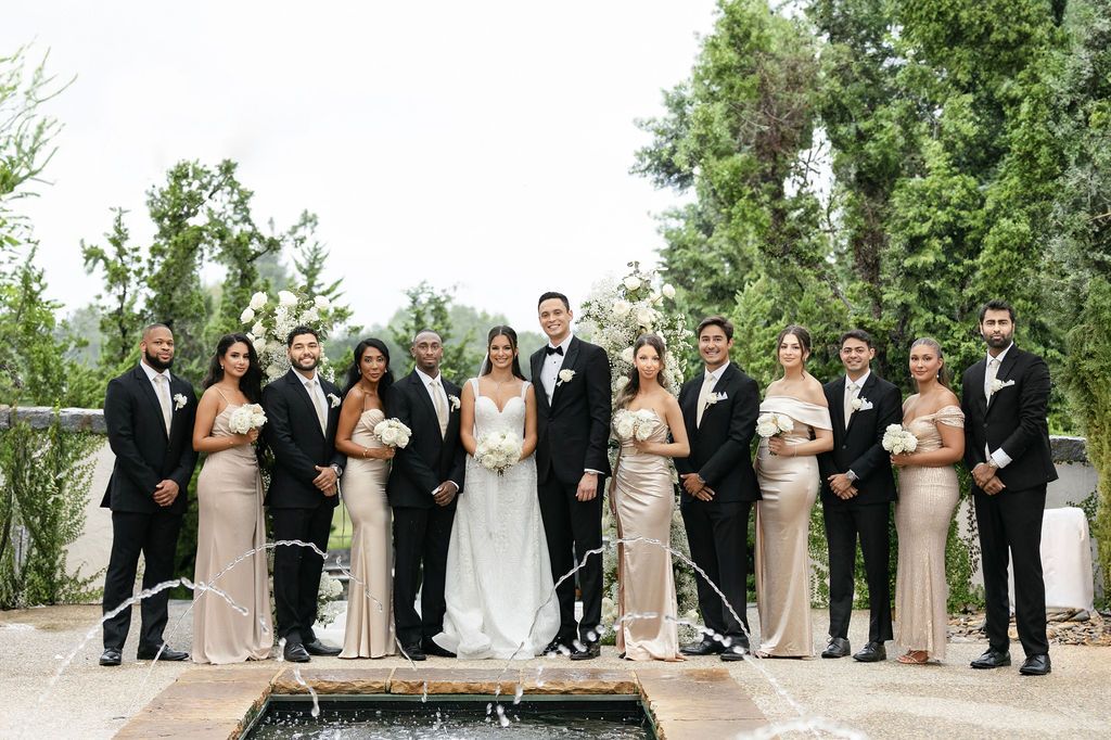 Wedding party poses on stone patio with bridal party in champagne dresses and groomsmen in black tuxedos.