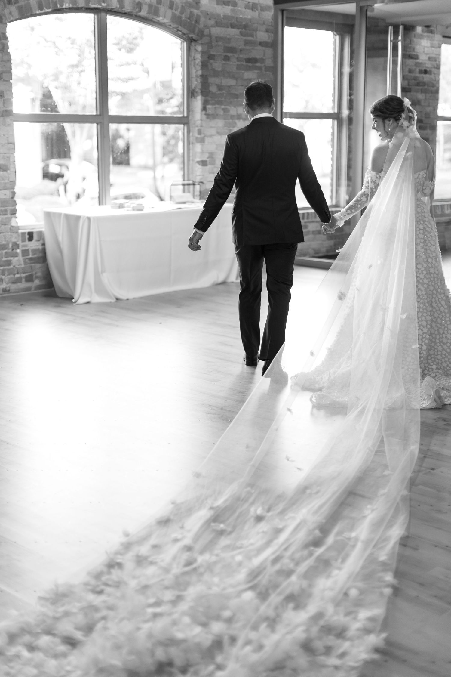 Black and white photo of bride and groom walking hand in hand, bride wearing long flowing veil trailing behind her.