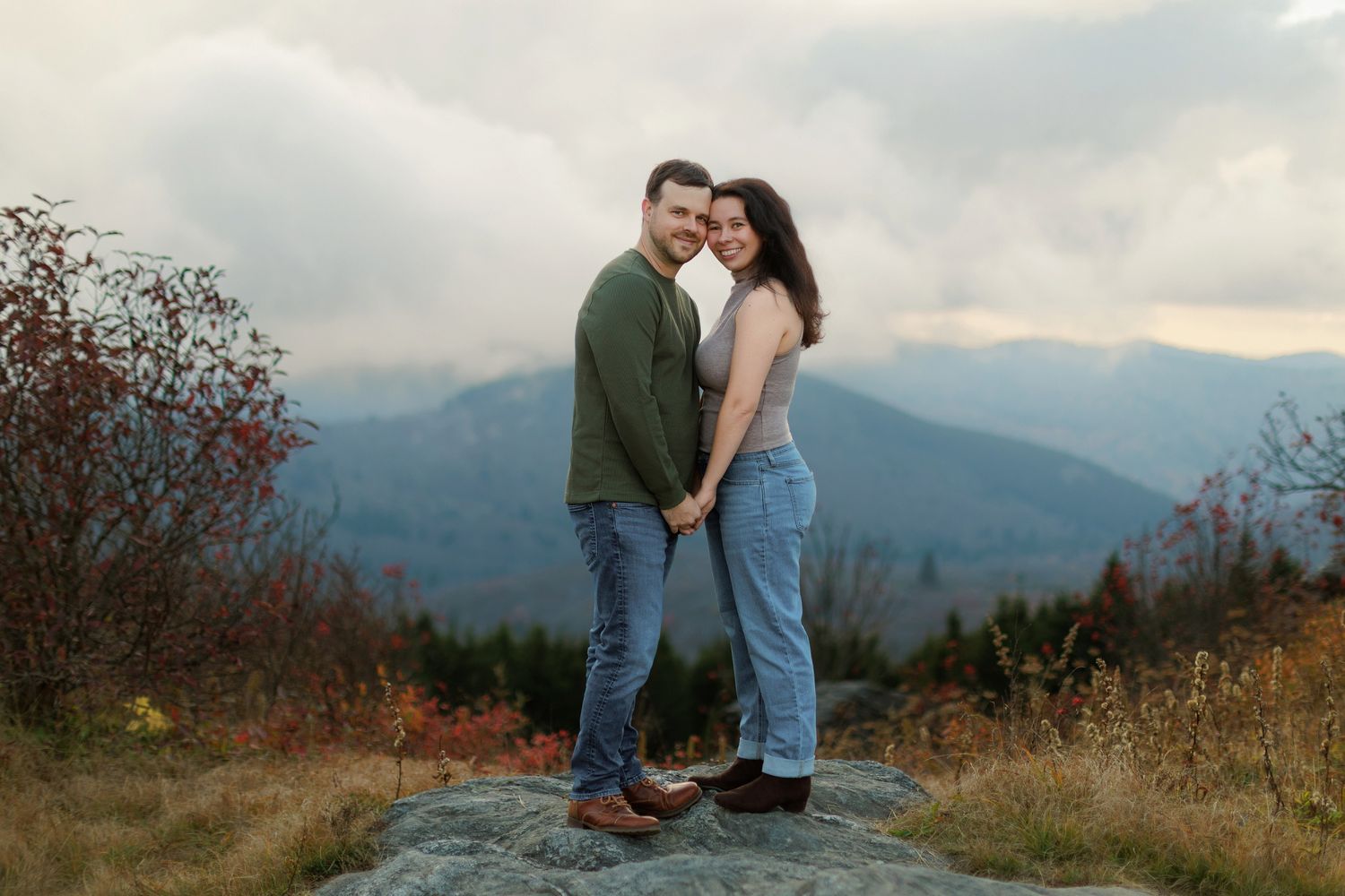 A couple shares a romantic moment on a hilltop overlooking misty mountains during sunset.