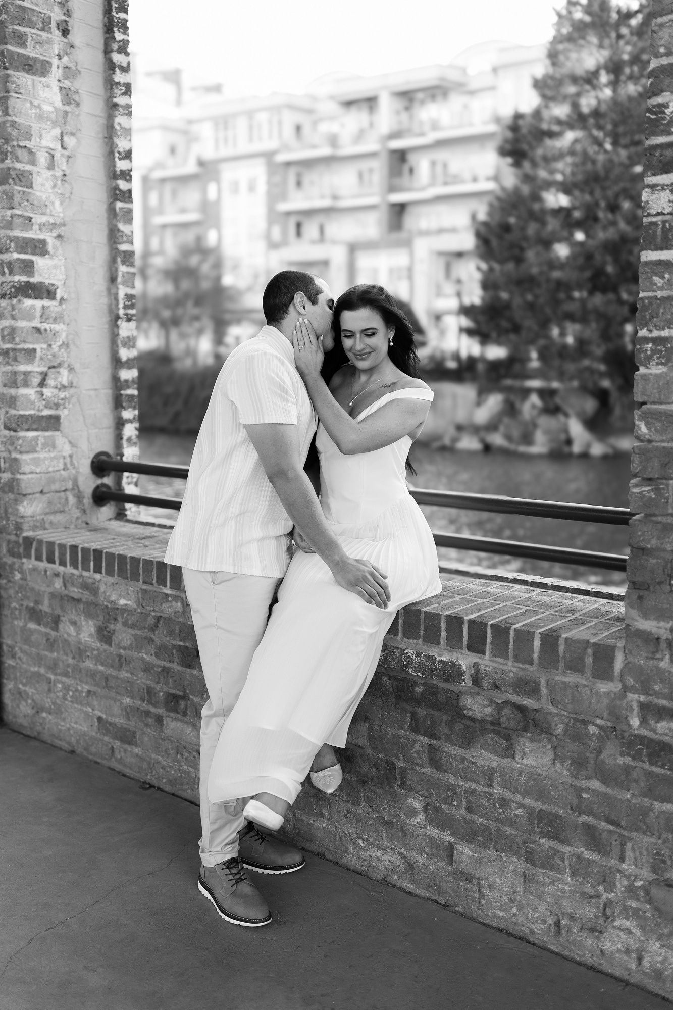 Black and white photo of a couple in white outfits posing playfully on a brick wall.