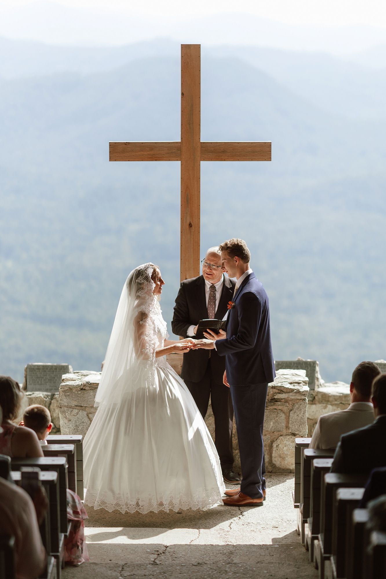 Bride and groom exchange vows before a wooden cross at an outdoor mountain wedding ceremony.
