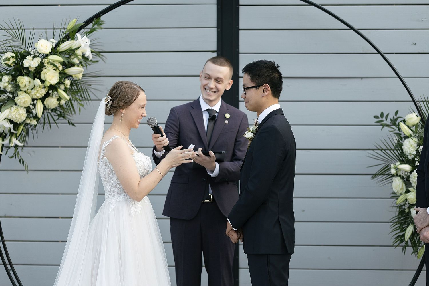 A wedding ceremony takes place under a floral arch against a gray wall with the officiant leading the service.