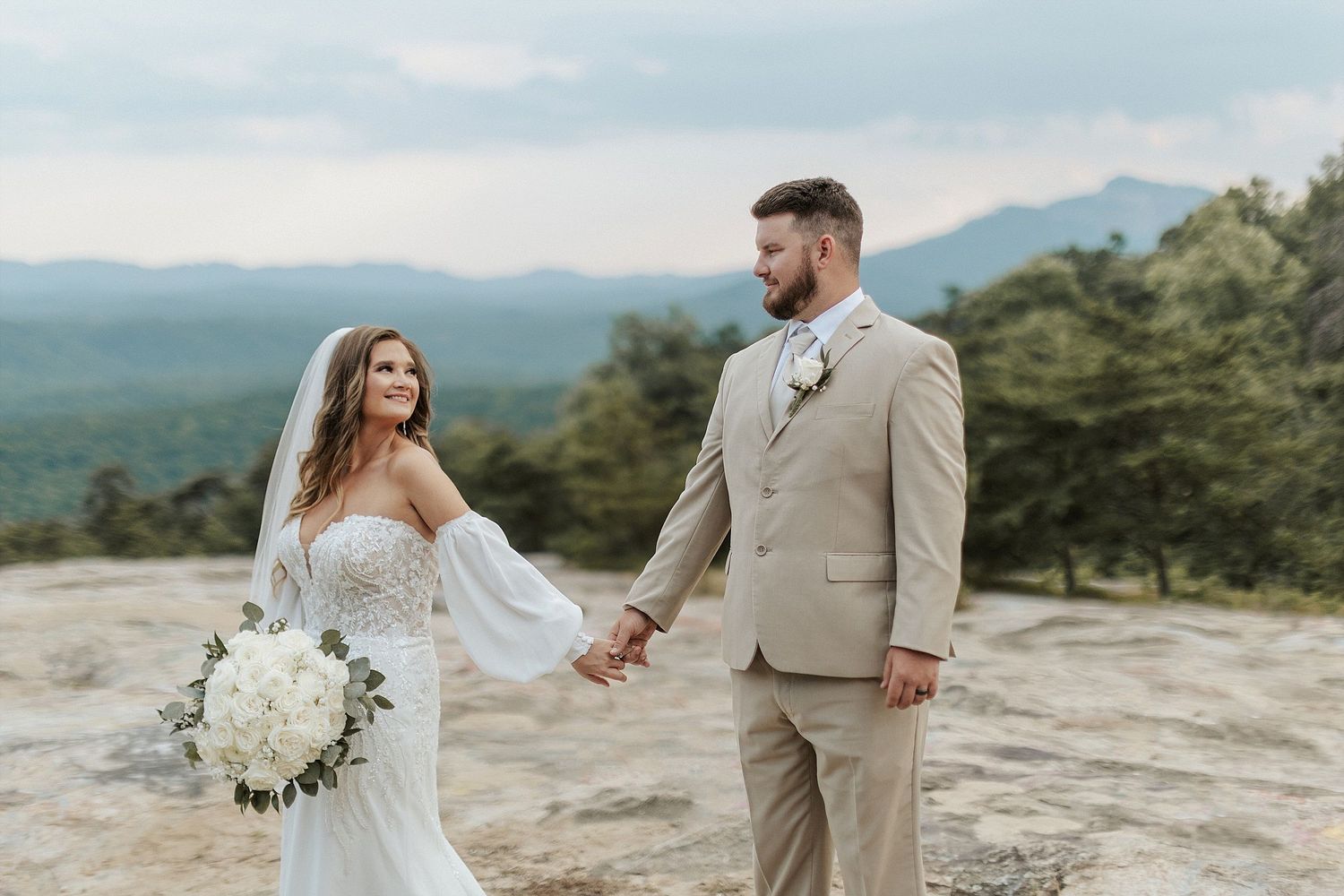 A wedding couple stands on a mountain overlook with scenic rolling hills in the background.