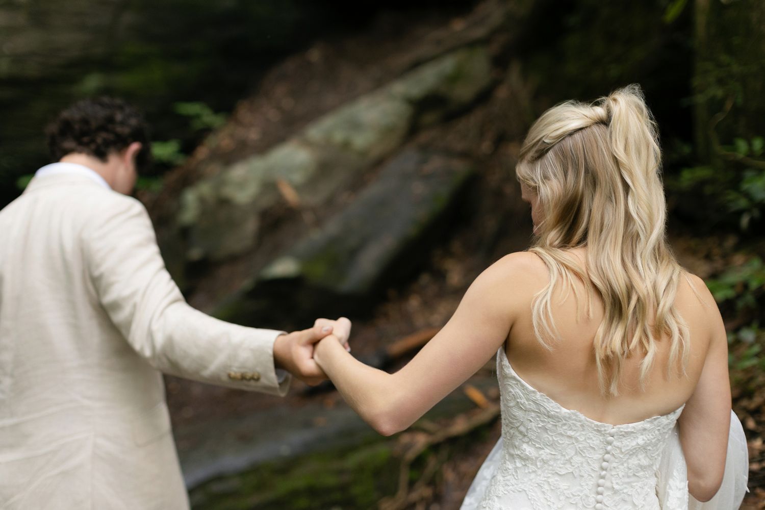 A couple holds hands against a blurred natural backdrop with a waterfall during an outdoor wedding ceremony.