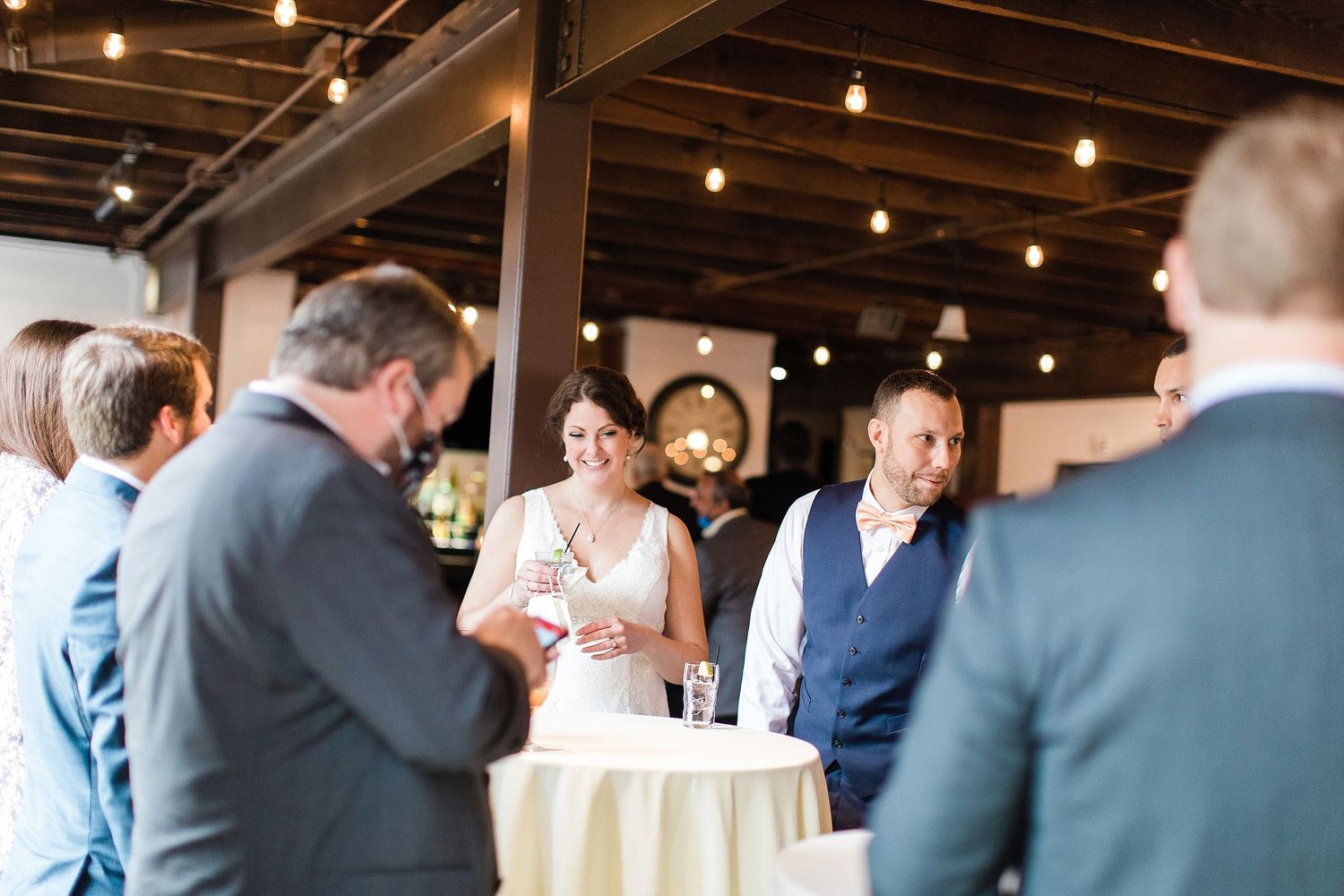 Wedding guests gather around a cocktail table for drinks and conversation in an industrial-style venue.
