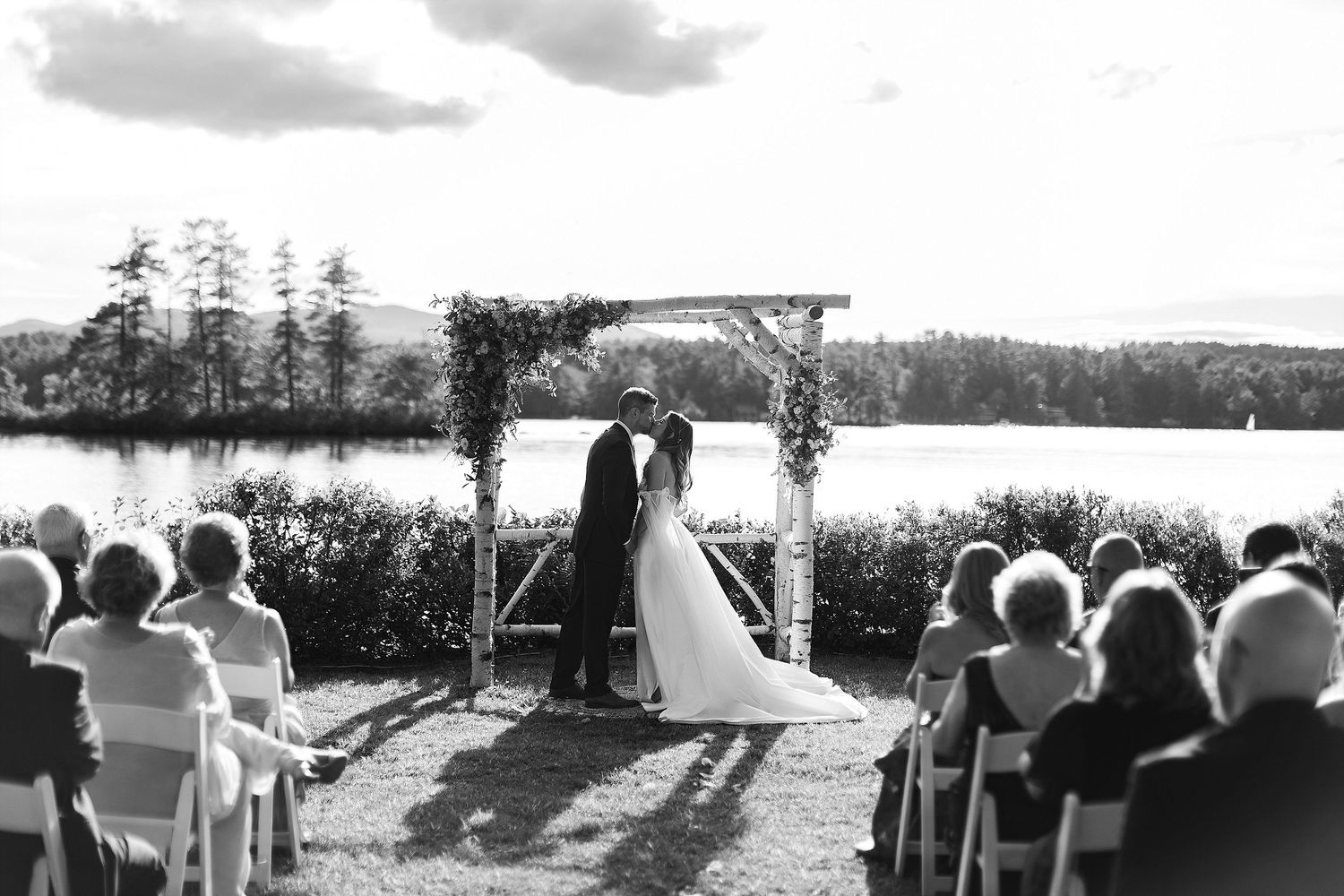 A black and white photo of a lakeside wedding ceremony with guests seated on white chairs.