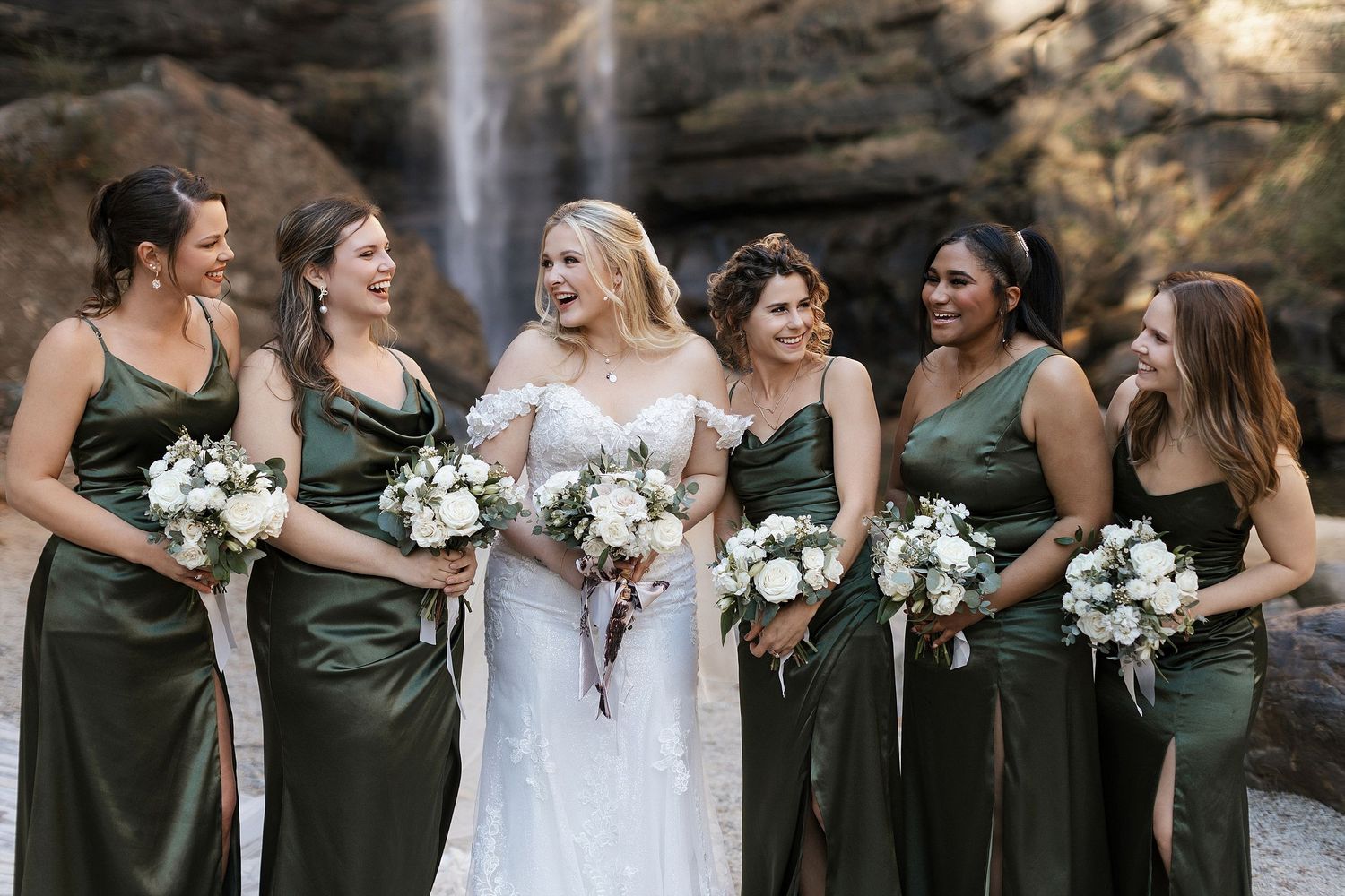 Wedding party in olive green bridesmaid dresses and white bridal gown pose with bouquets in front of majestic waterfall.