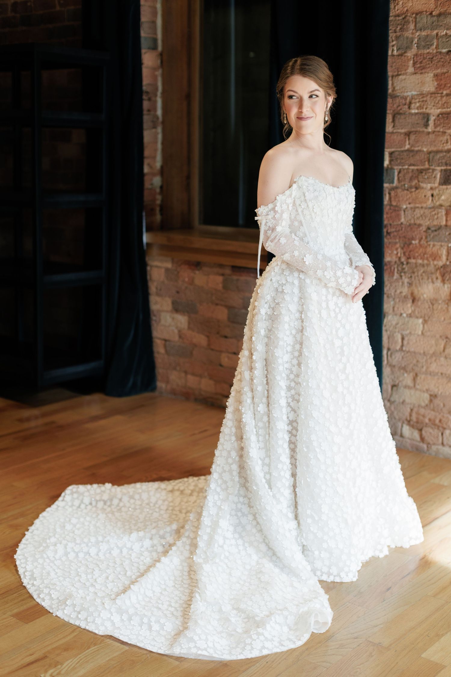 Bride in elegant off-shoulder white ball gown with floral texture and long train, posing in rustic brick venue.