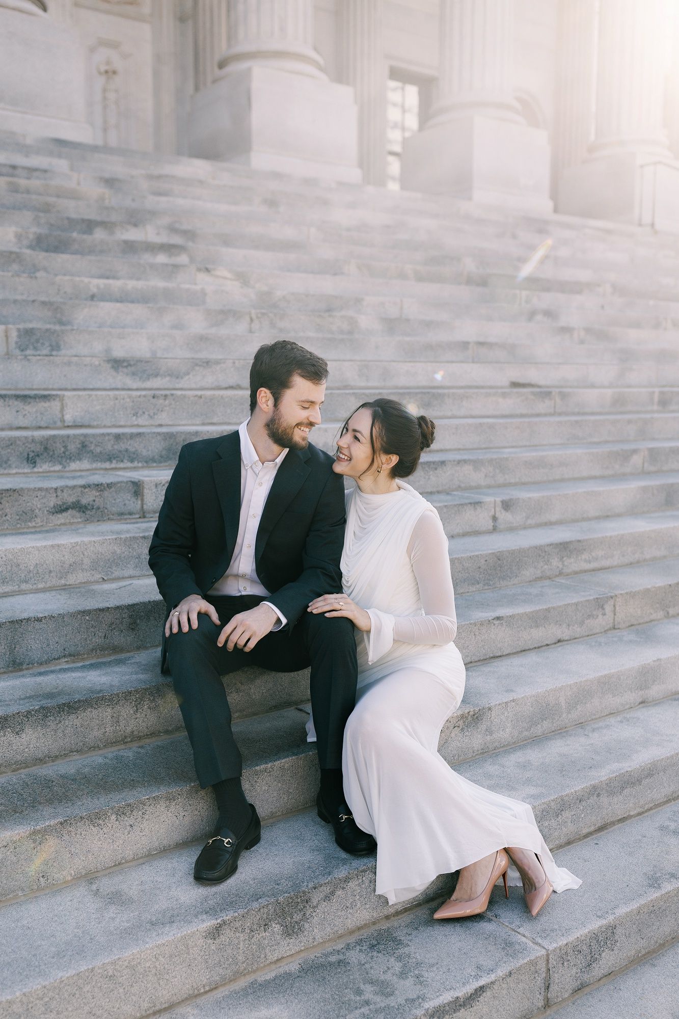 A couple in formal attire share intimate moments while sitting on outdoor stone steps in black and white tones.