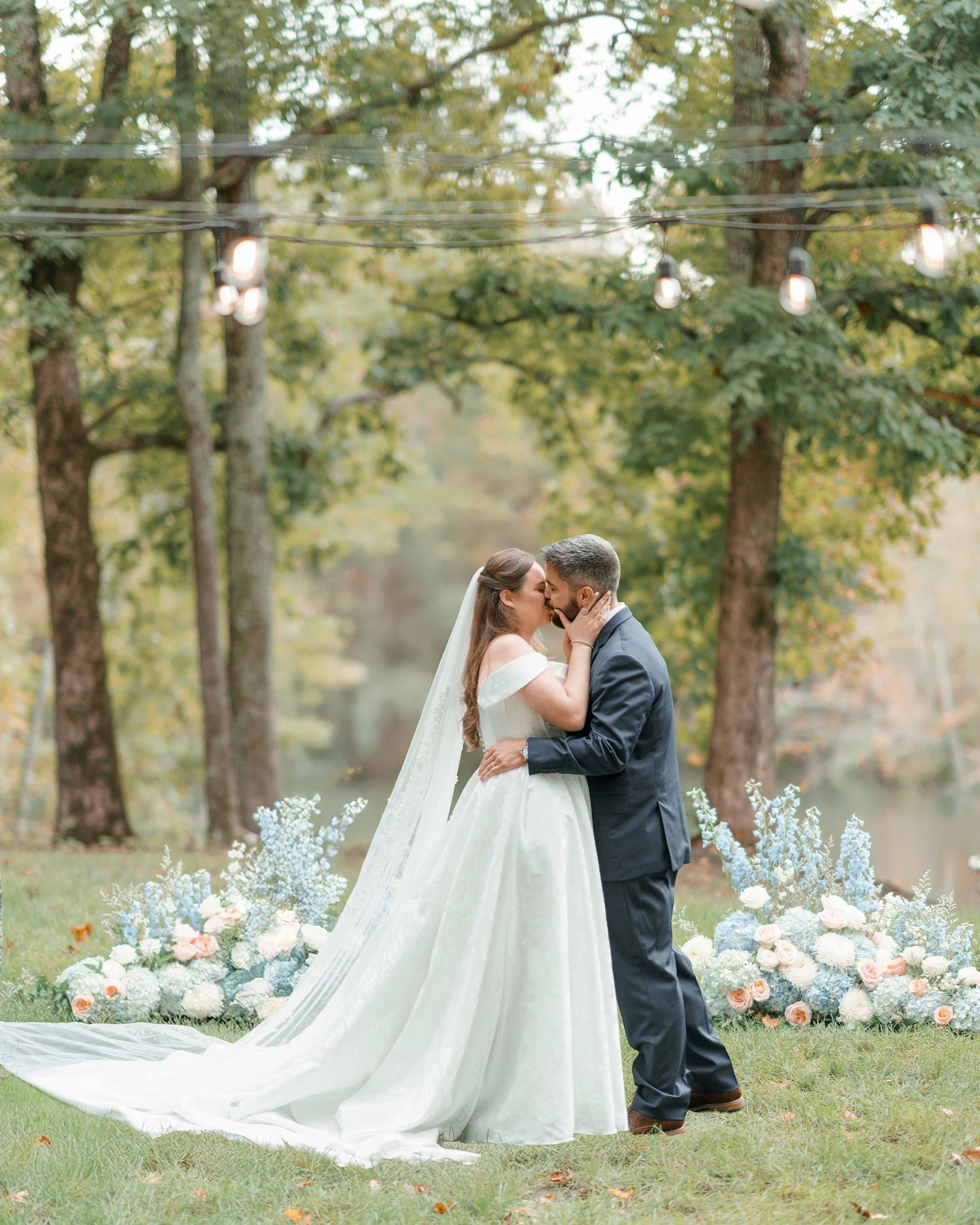 Bride and groom share first kiss at outdoor wedding with pastel floral backdrop and string lights.