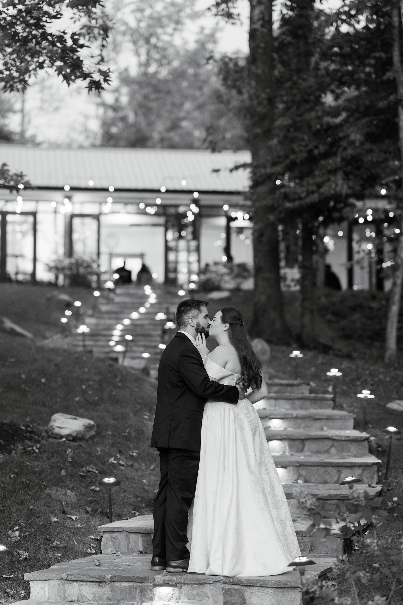 Bride and groom share a romantic kiss on candlelit stone steps outside their wedding venue at dusk.