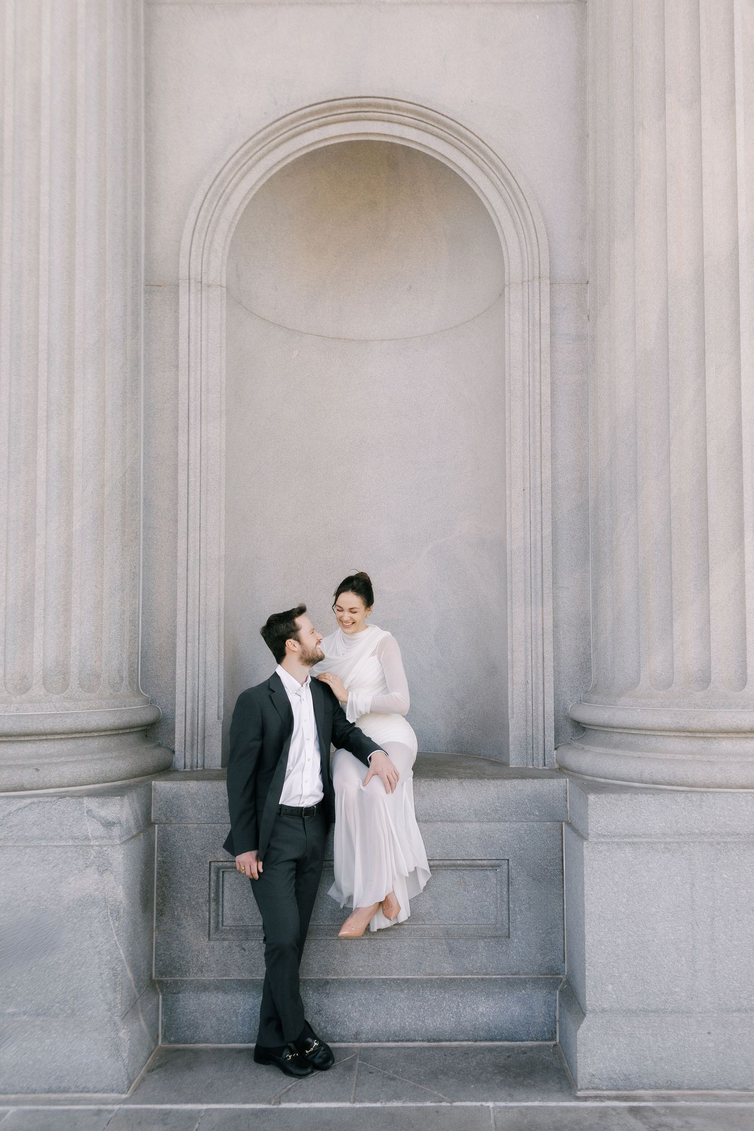 Elegant couple in formal attire posing in a classical stone archway, man in dark suit, woman in flowing white dress.