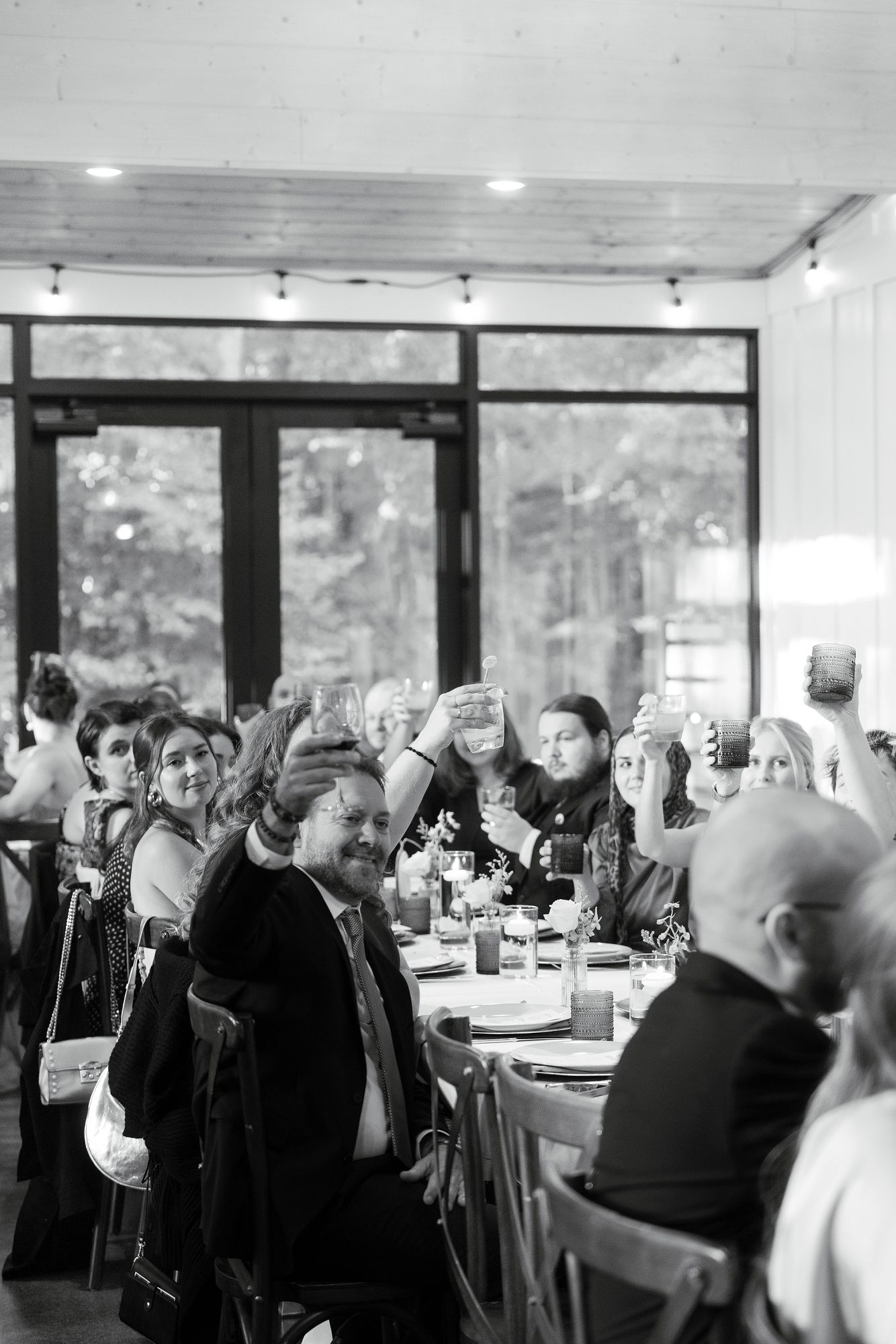Black and white photo of wedding reception guests raising glasses for a toast inside an elegant venue.